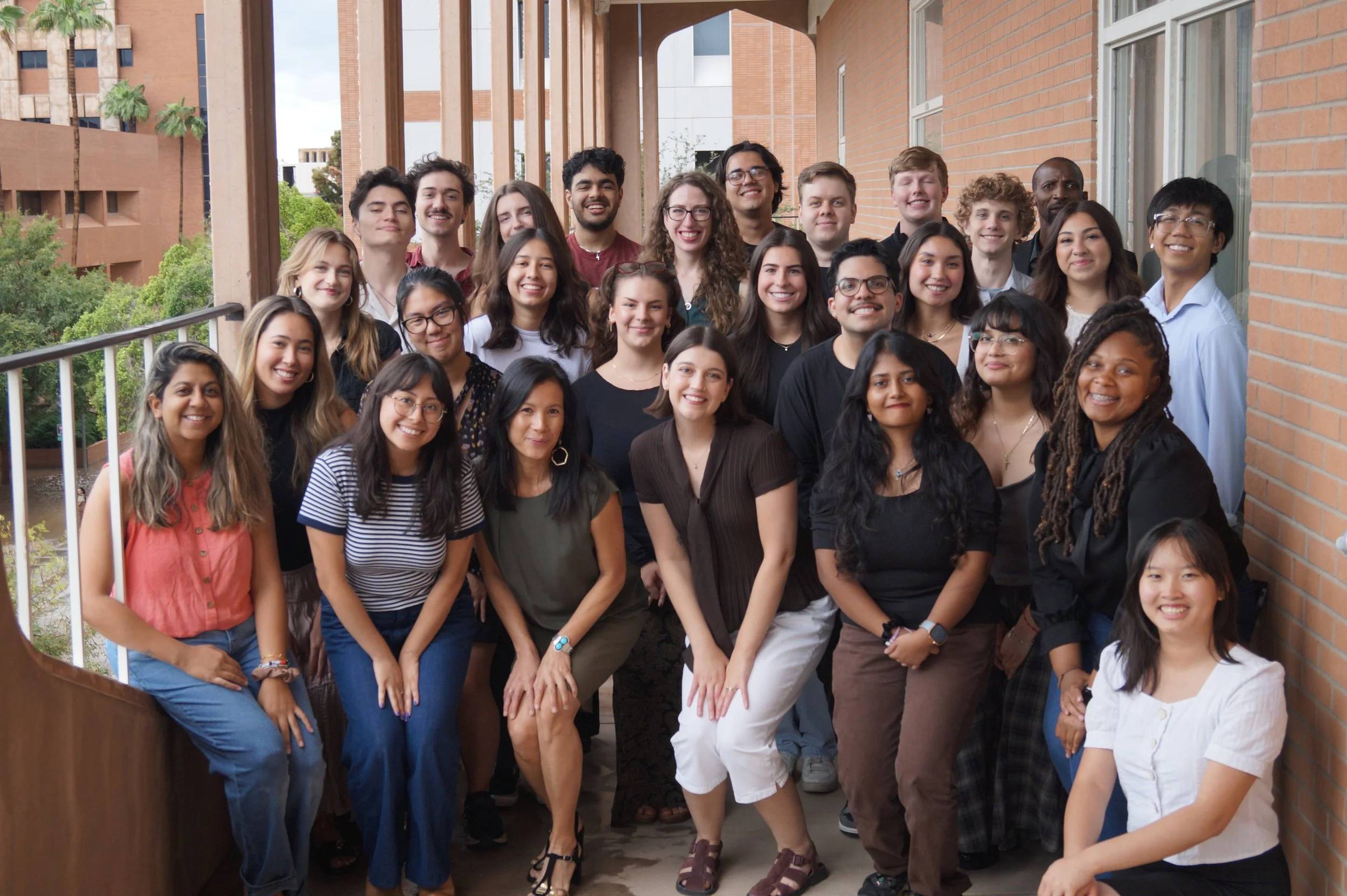 Group of young adults posing on a balcony outdoors with trees and modern buildings in the background.