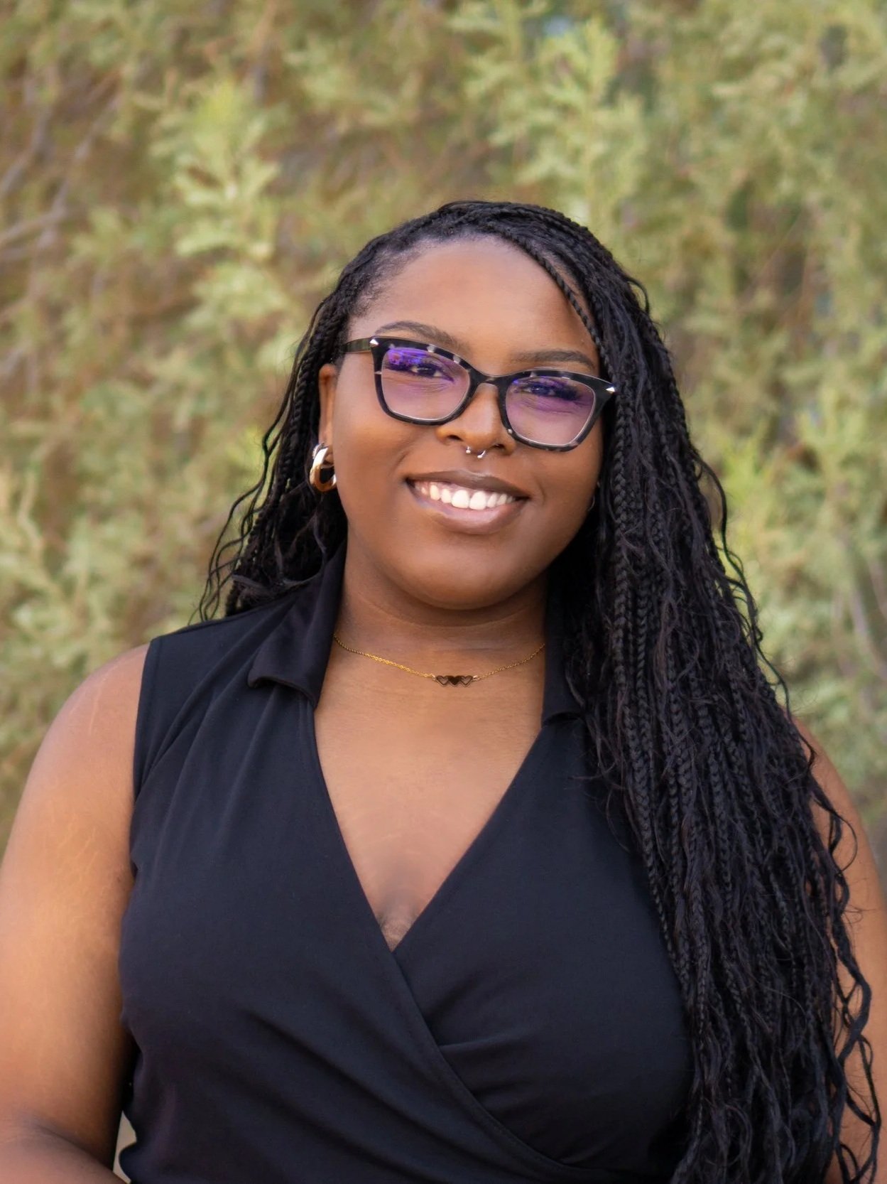 A woman with glasses, hoop earrings, a nose ring, and a necklace, smiling outdoors with trees in the background.
