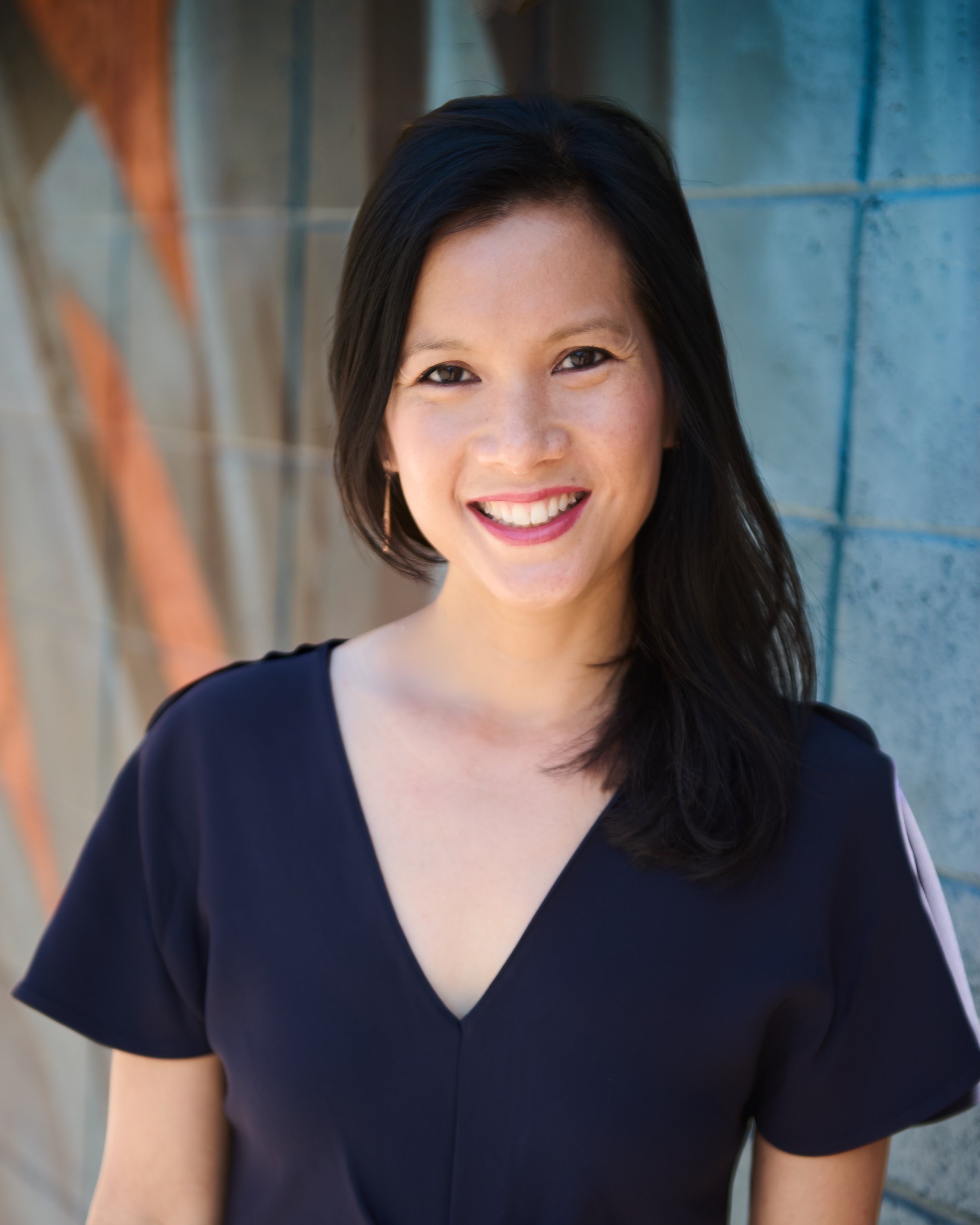 A young woman with long dark hair smiling outdoors with trees in the background.