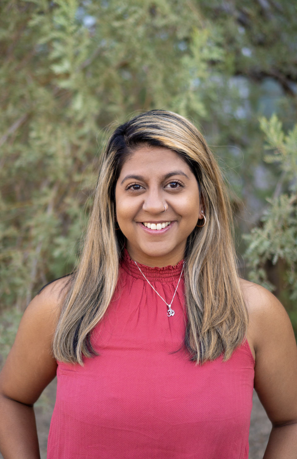A smiling woman with light brown hair with blonde highlights, wearing a red sleeveless top, standing outdoors with greenery in the background.