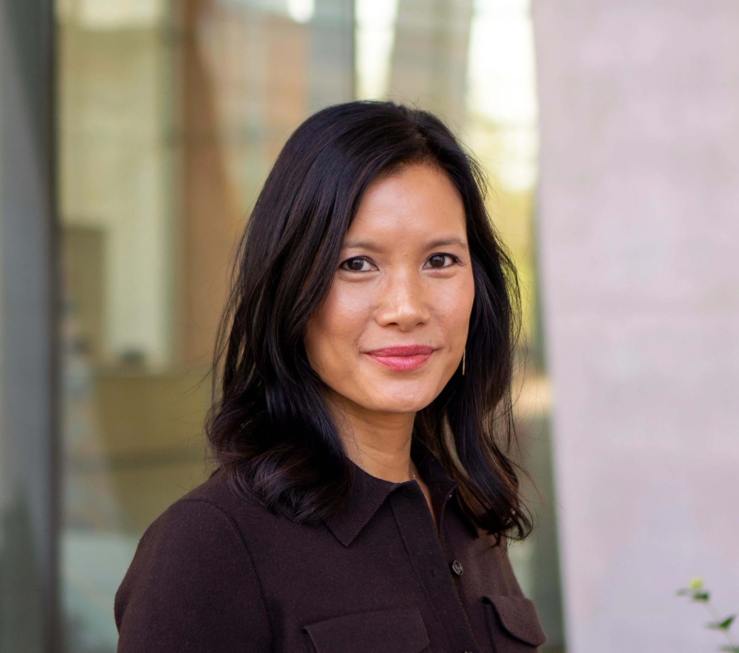 A young woman with long dark hair smiling outdoors with trees in the background.