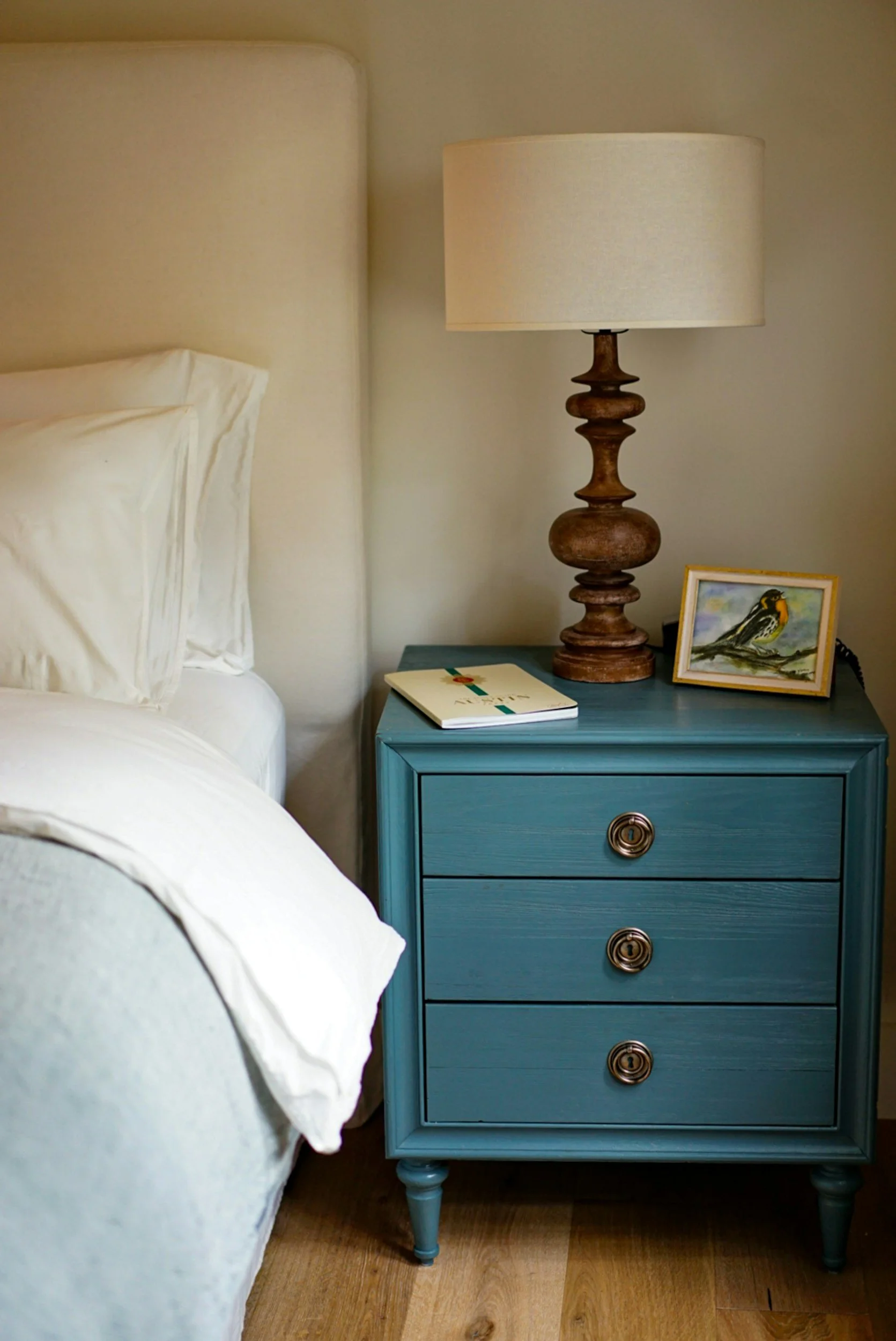 A blue bedside table with three drawers, a lamp with a beige shade, a framed bird painting, and a white book, next to a bed with white bedding and pillows.