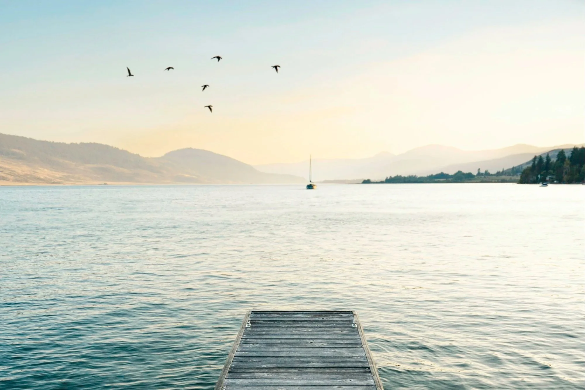 A tranquil lake scene at sunset with mountains in the background, a wooden dock extending into the water, a sailboat in the distance, and birds flying overhead.
