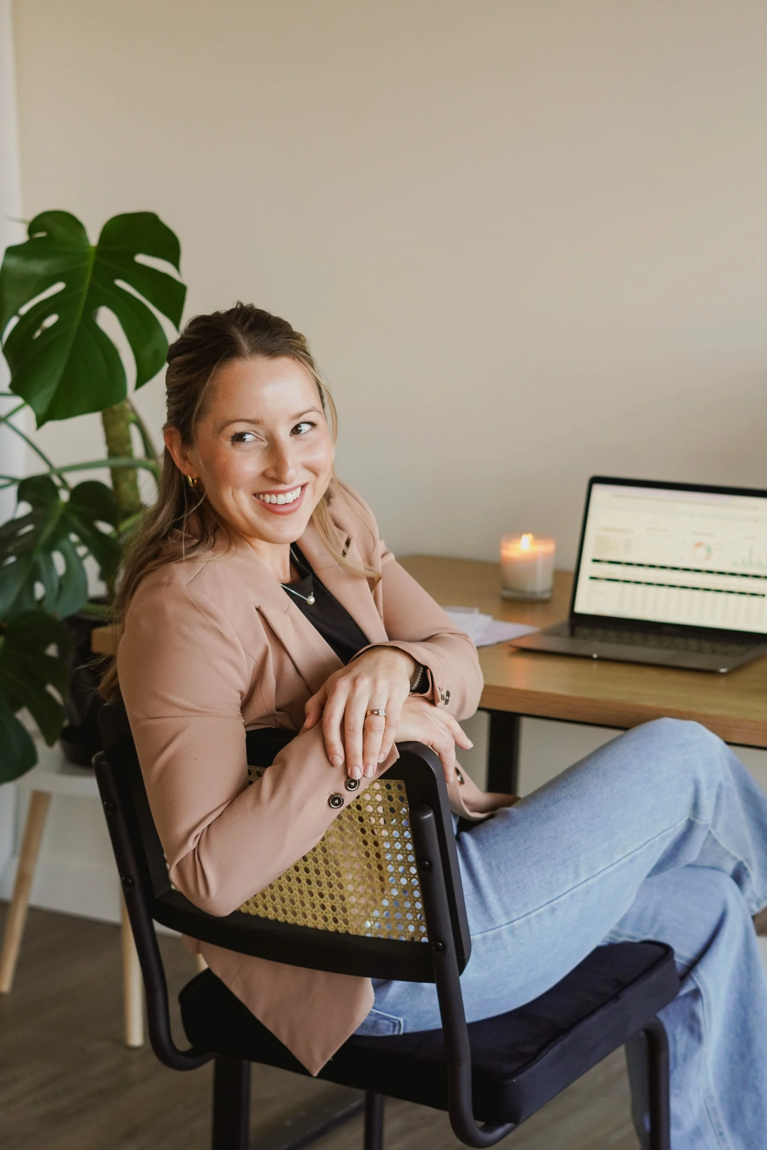 A woman with light brown hair, smiling, sitting on a black chair in an office. She wears a beige blazer, black shirt, and light blue jeans. Behind her is a wooden desk with a laptop displaying graphs, a lit candle, and some papers. There is also a large green plant to her left.