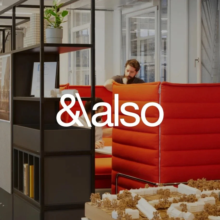 A man working in a modern office with orange high-backed booths and a black shelving unit in the foreground, large windows in the background, and a model of a building or city layout on a table.