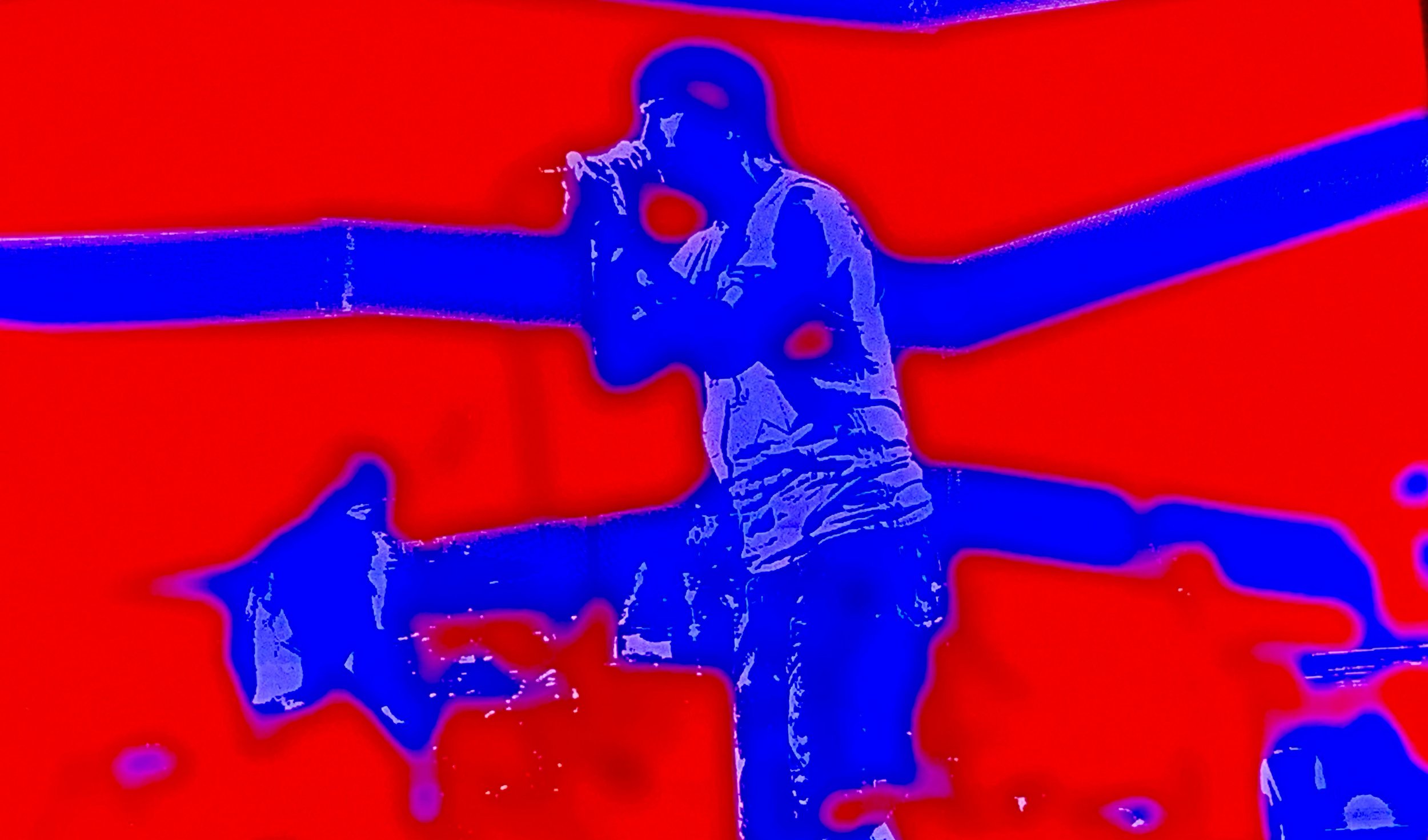 A person holding a microphone standing on stage with a red background, outlined in blue and purple, in a stylized, artistic filter.