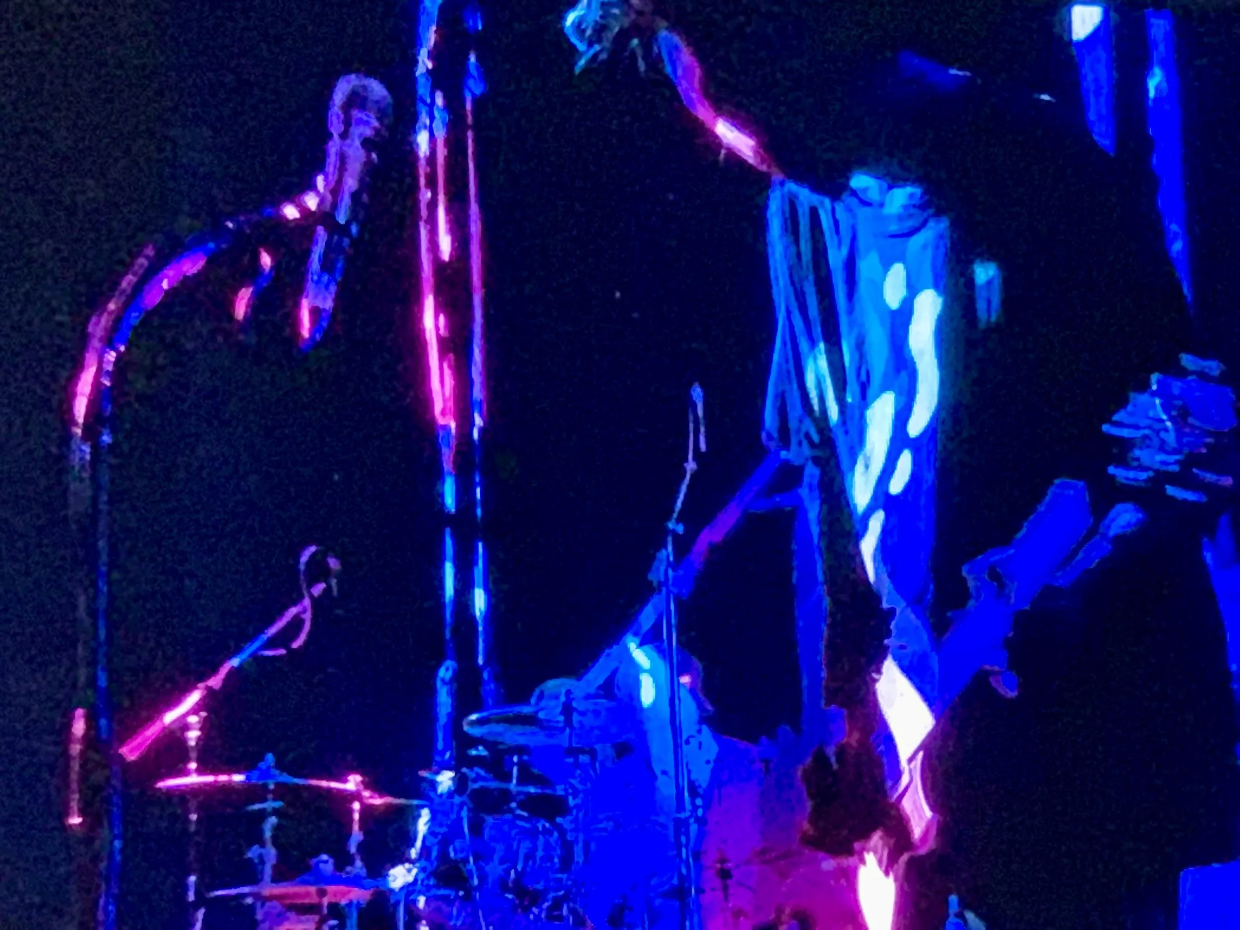 Two musicians illuminated with neon pink and blue lights, one playing drums and the other playing guitar, on a dark stage.