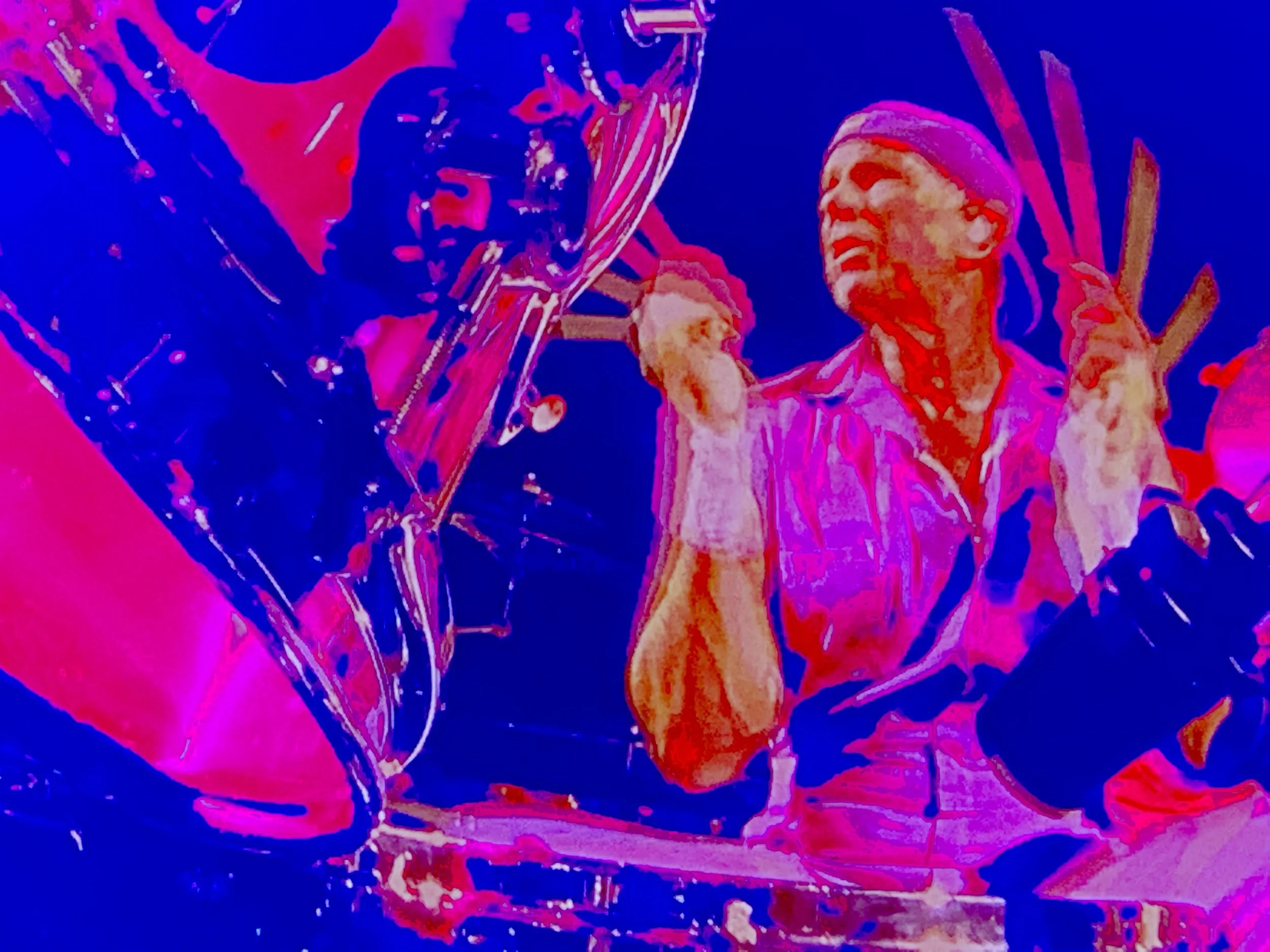 A vibrant, stylized photo of two musicians performing on stage. One person is playing an electric guitar and wearing a pink shirt and pink bandana, smiling and enjoying the music, with a dark background.