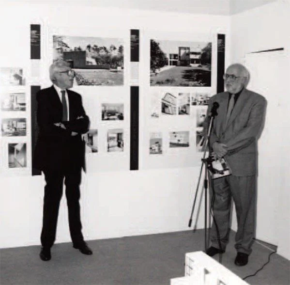 Two older men in suits standing in an art gallery or exhibition space, observing architectural or real estate photographs displayed on the wall, with one man speaking into a microphone.
