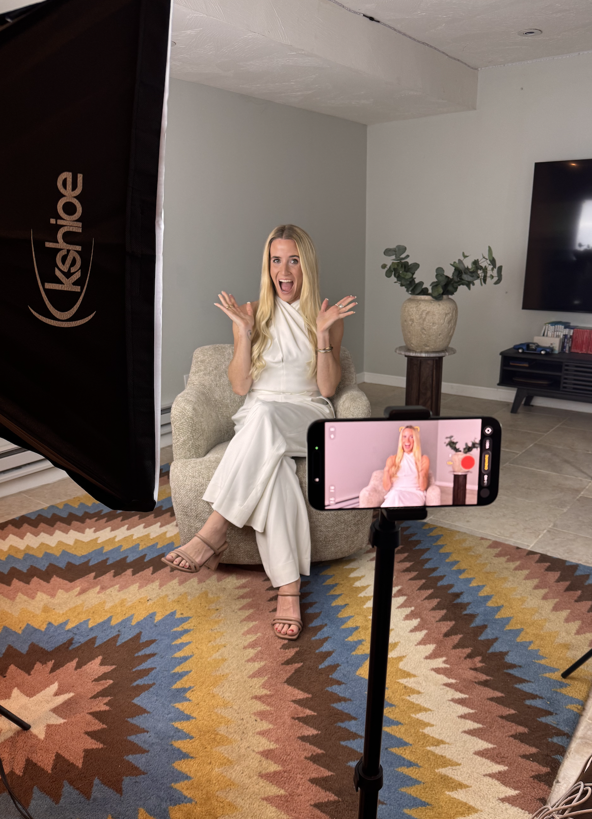 A woman in a white outfit sitting on a beige armchair, smiling and gesturing with excitement, during a video recording in a living room with a colorful rug, a potted plant, and a flat-screen TV.
