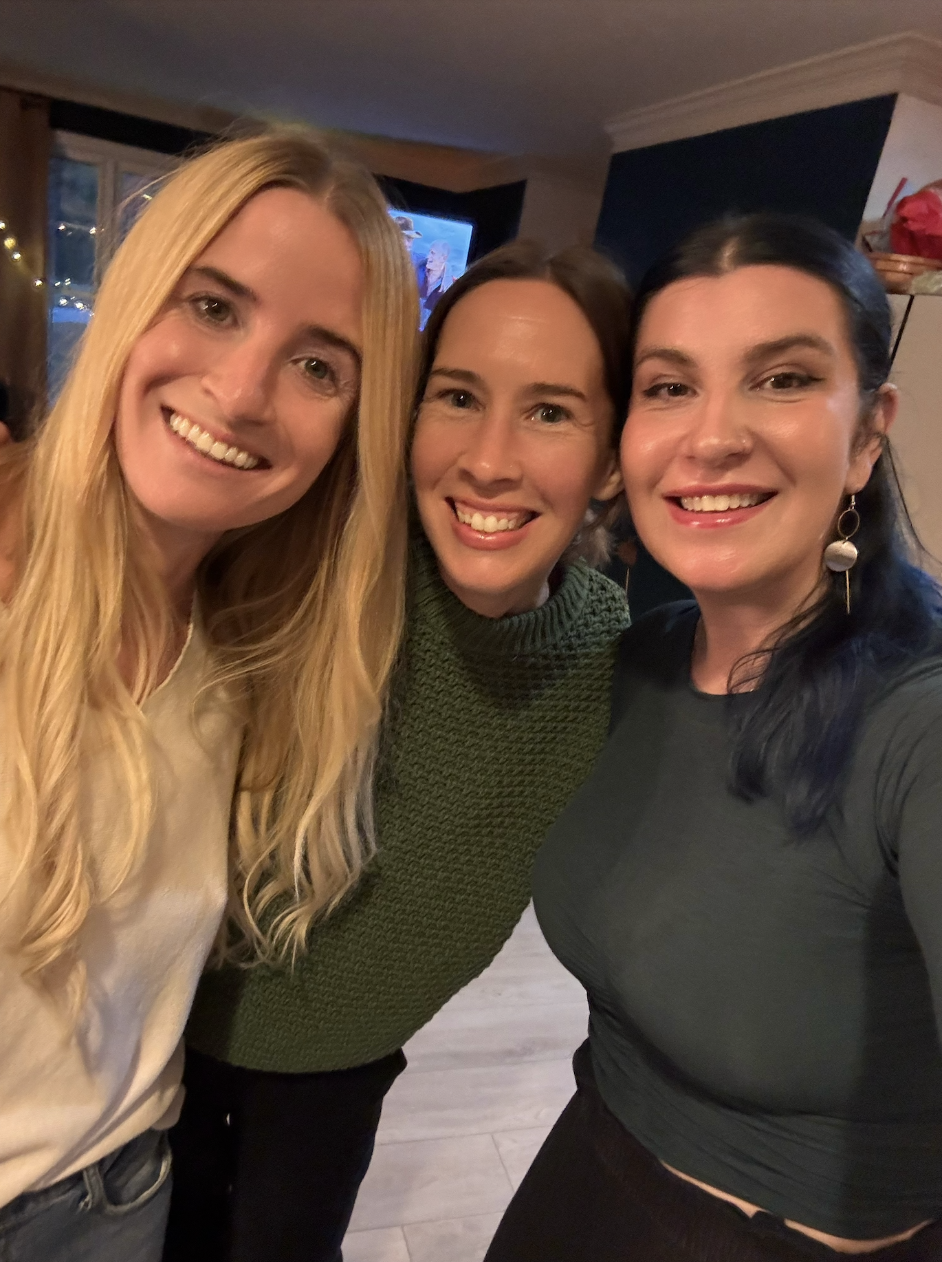 Three smiling women taking a selfie in a cozy room with Christmas lights and a television in the background.
