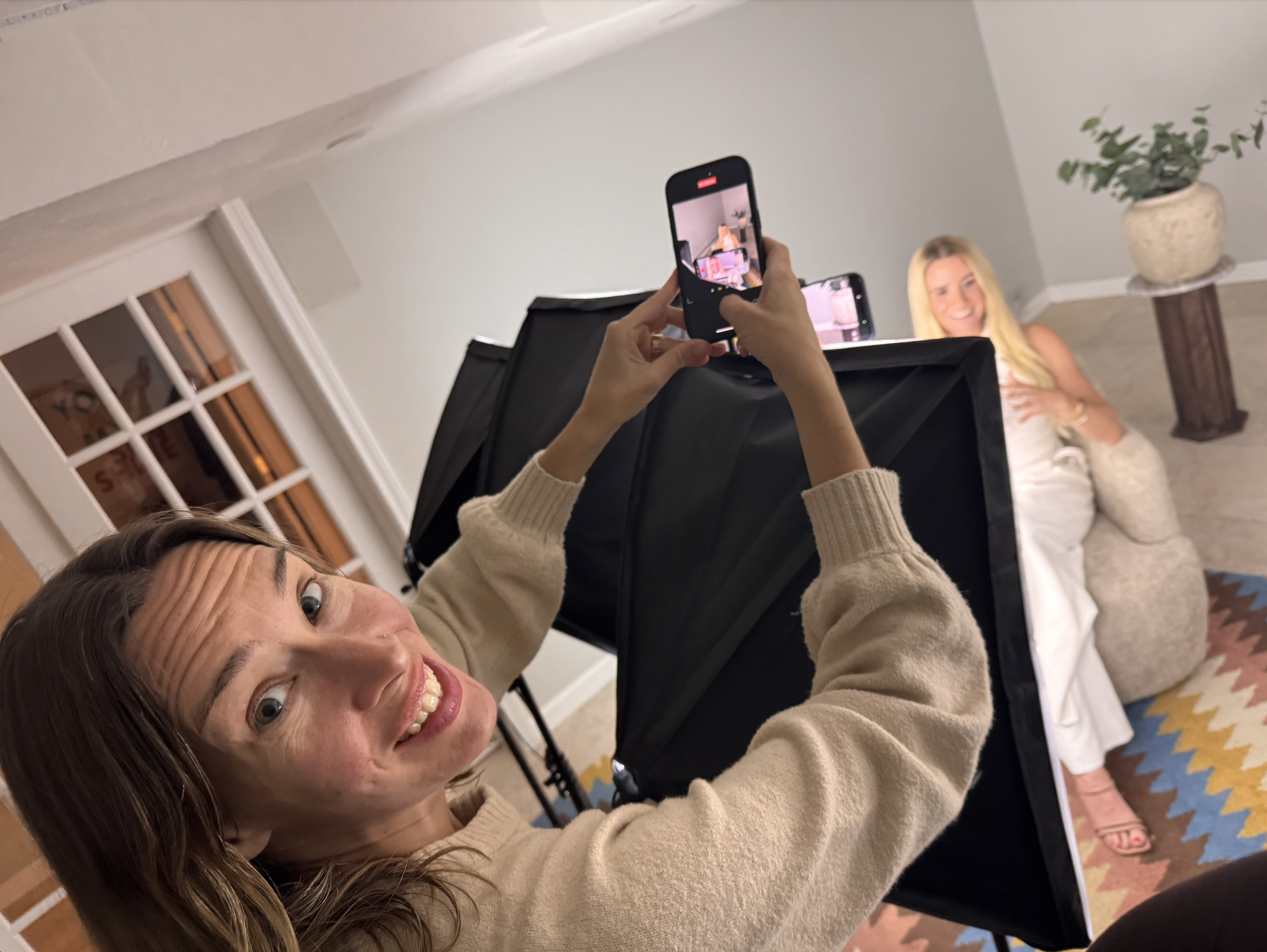 A woman with brown hair and glasses takes a selfie while smiling, with a background of a woman with long blonde hair posing in front of a camera set up and a houseplant in a pot, in a room with a patterned rug.