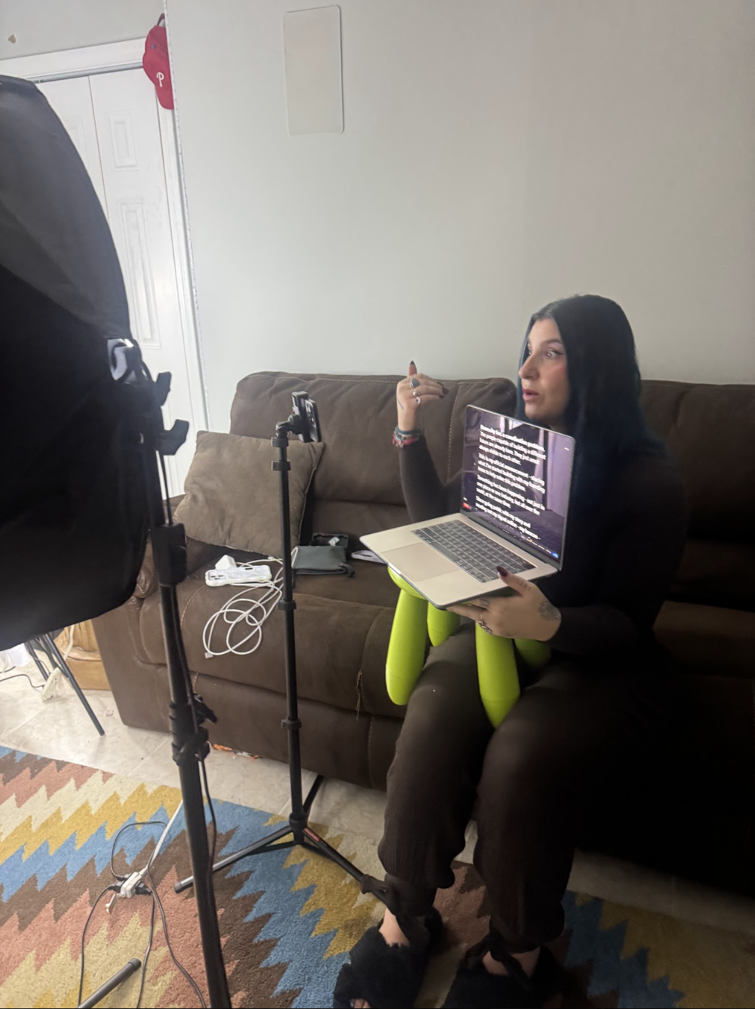 Woman with dark hair sitting on a brown couch, holding a laptop with text on the screen, gesturing with her hand, in a room with a colorful rug, and filming equipment in front of her.