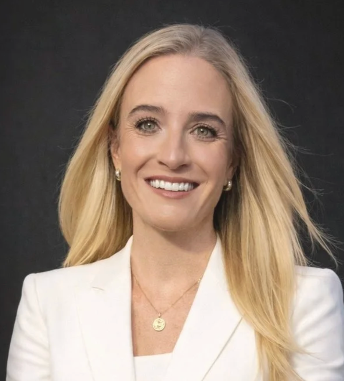 A smiling woman with long blonde hair, wearing a white blazer and a gold necklace with a circular pendant, against a black background.