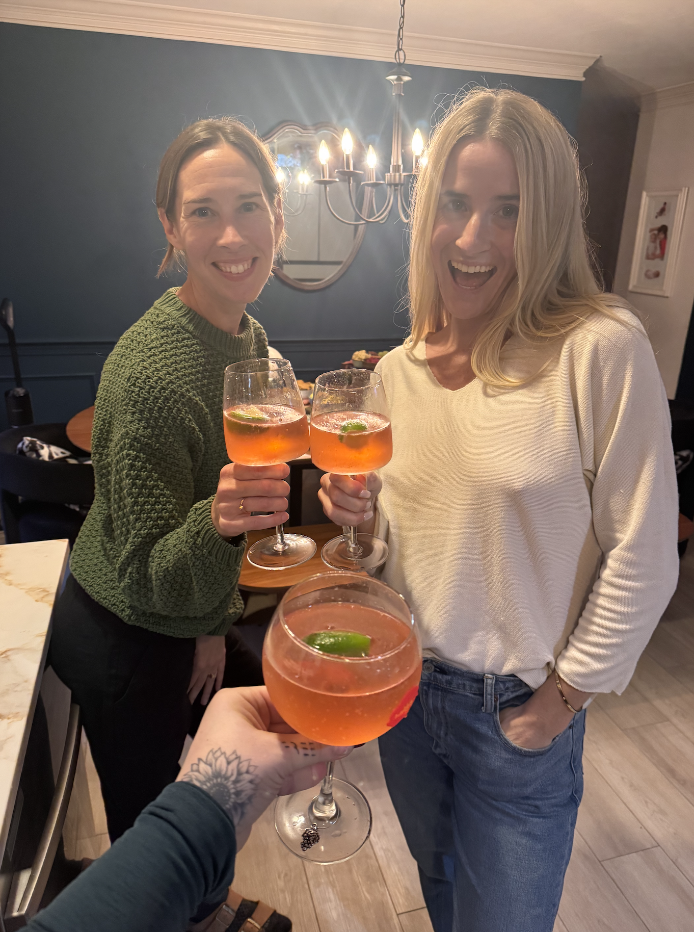 Two women smiling and holding glasses of a pink cocktail with lime slices, celebrating together in a warmly lit room with a chandelier hanging above.