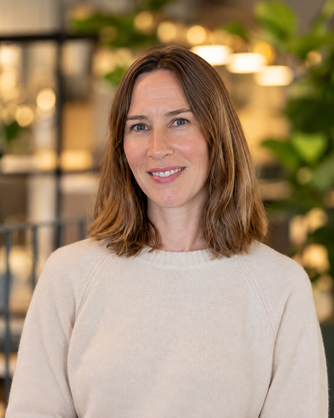 A woman with shoulder-length brown hair and blue eyes, smiling, wearing a cream-colored sweater in an indoor setting with blurred warm lights and greenery in the background.
