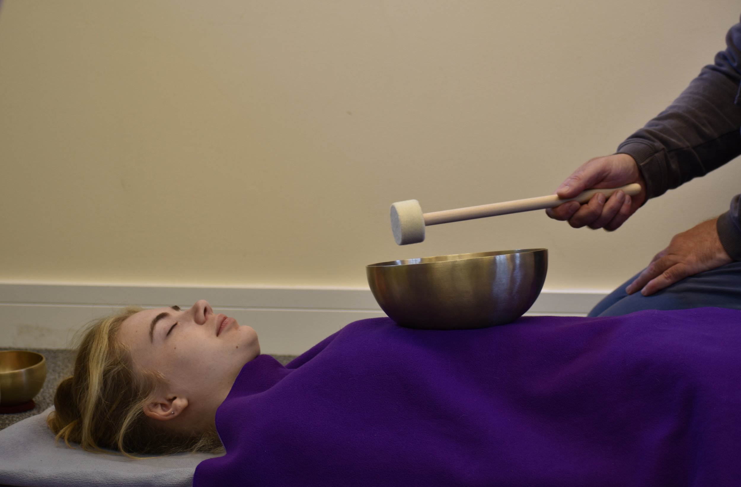 Person lying on an orange mat receiving sound therapy from another person using a mallet and singing bowl.
