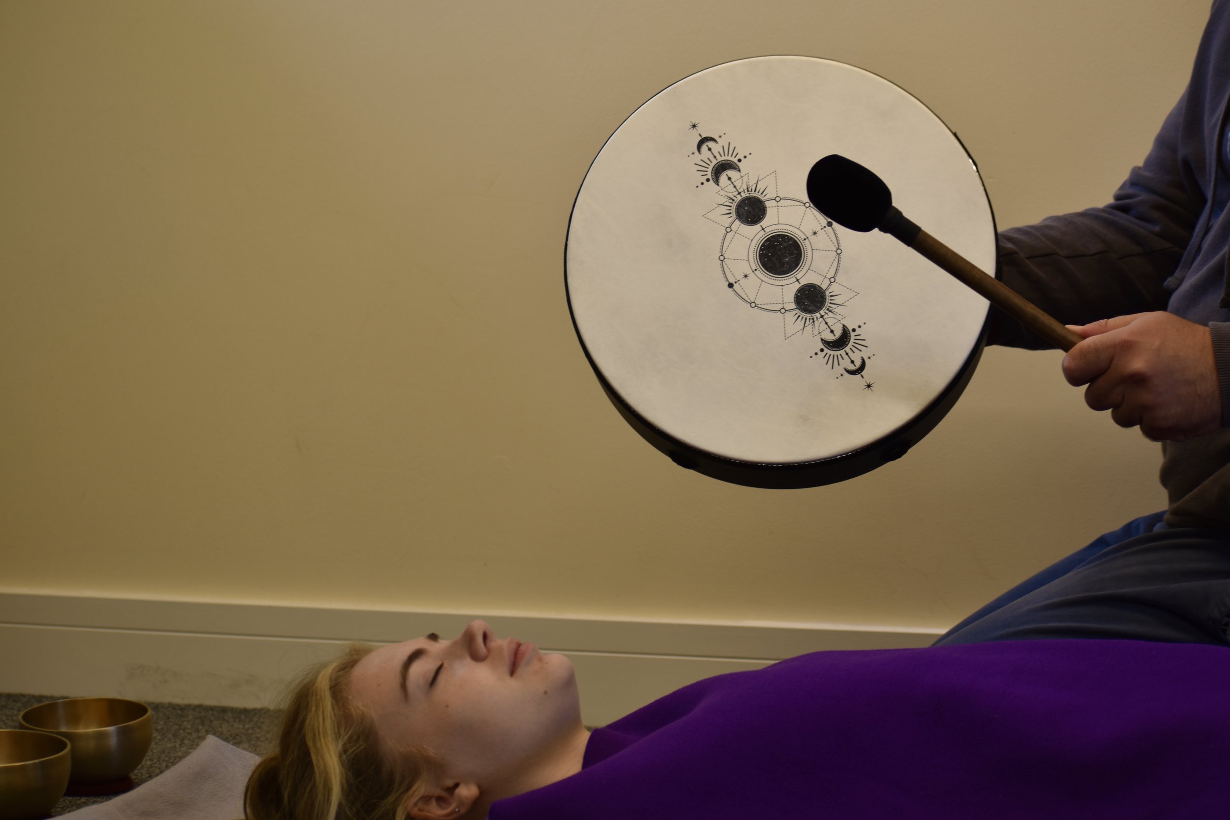 A person lying on their back with closed eyes, receiving sound therapy with a large hand-held drum decorated with celestial symbols held by another person.