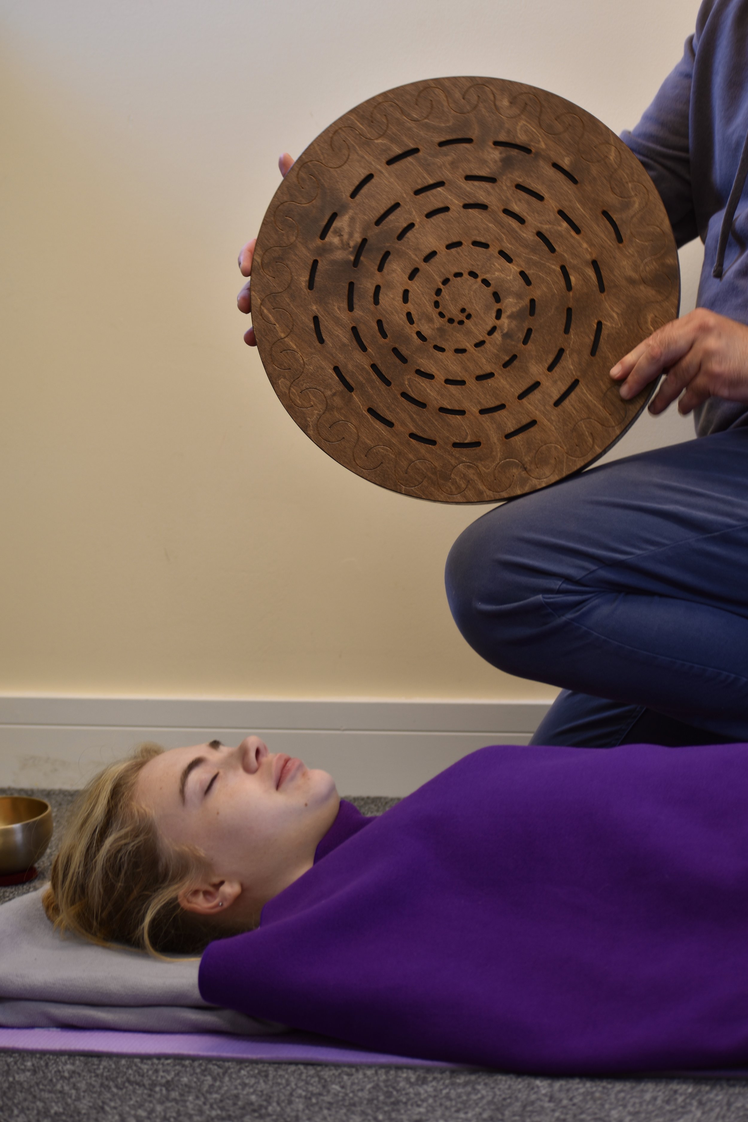 A woman lying on a yoga mat with eyes closed, while a person holds a wooden sound bowl above her for sound therapy.