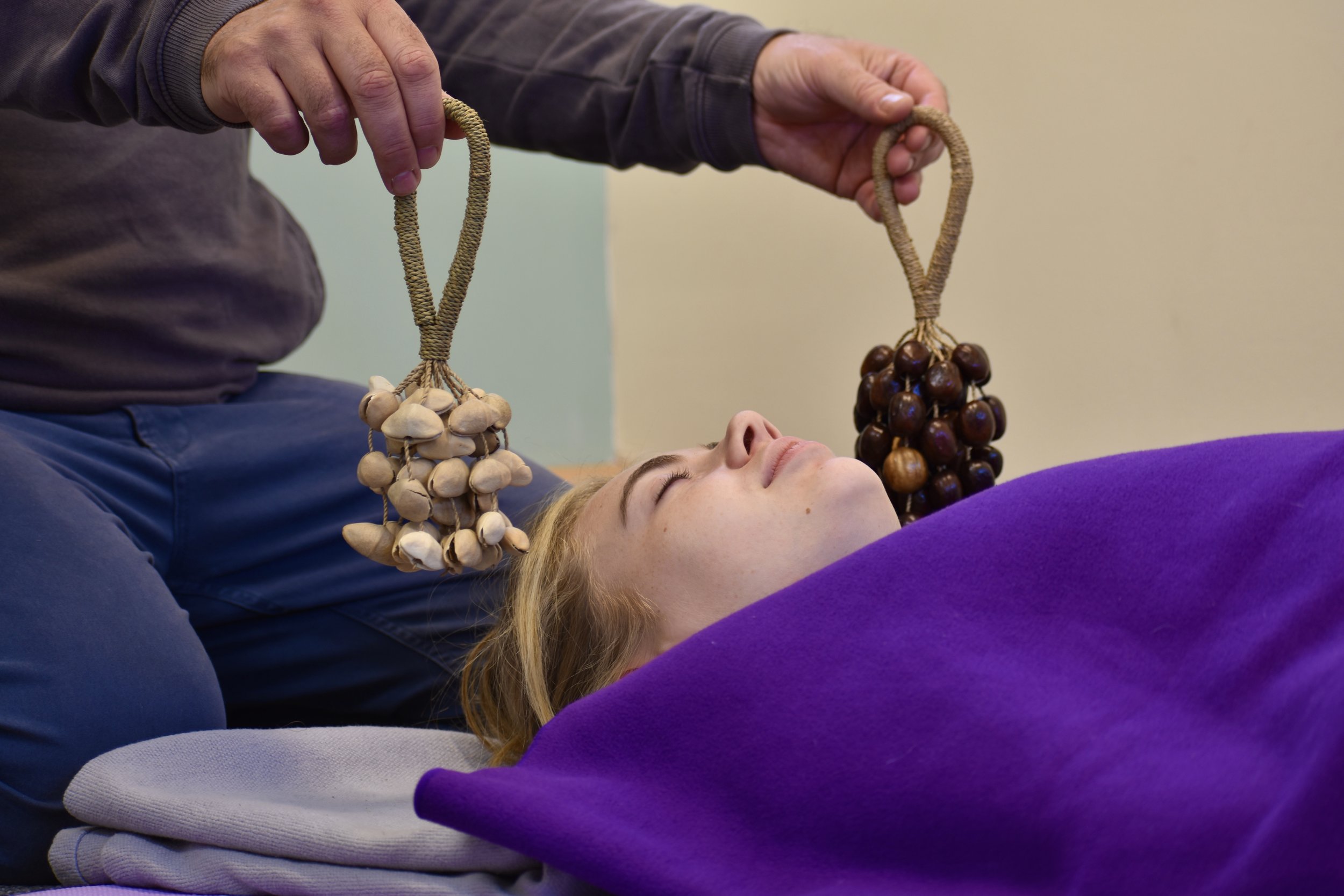 A woman lying down with a purple blanket over her chest, receiving a sound therapy treatment with two handheld instruments made of natural materials, held by a practitioner.
