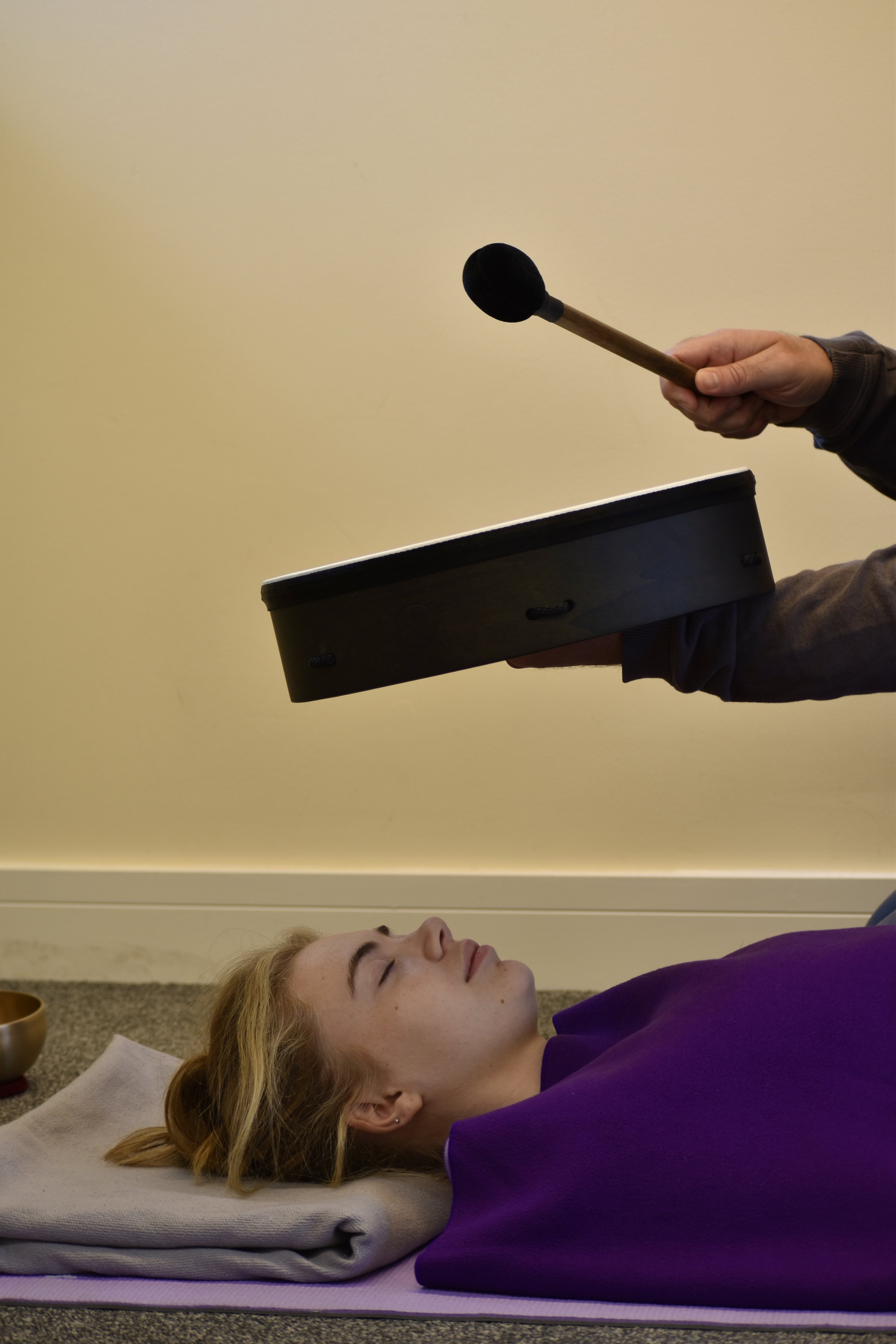 A woman lying on yoga mat with eyes closed, receiving sound therapy from a practitioner holding a gong and mallet.