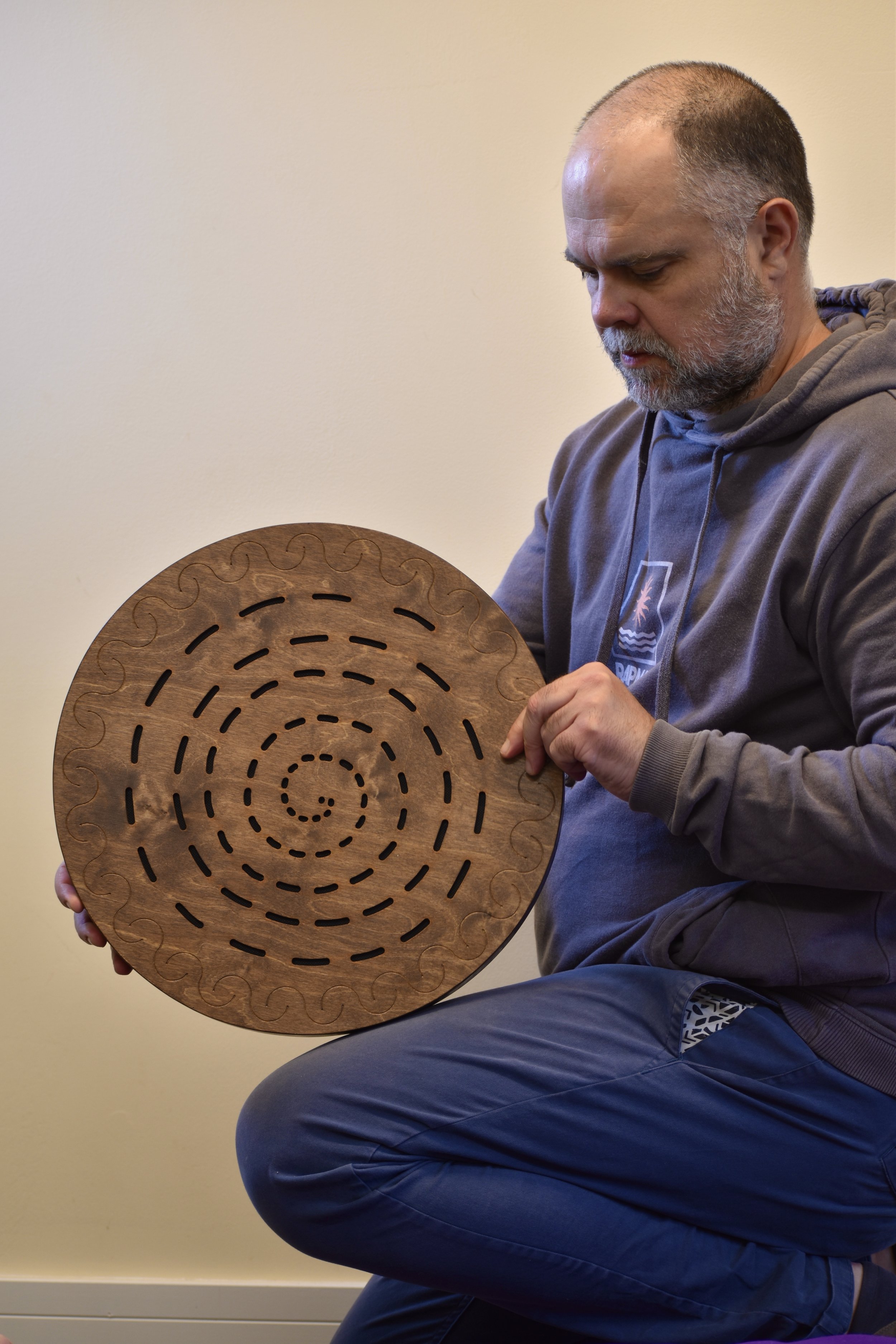 A man with a beard and no hair, dressed in a gray hoodie, is sitting and holding a circular wooden object with spiral and wave-like cutouts.