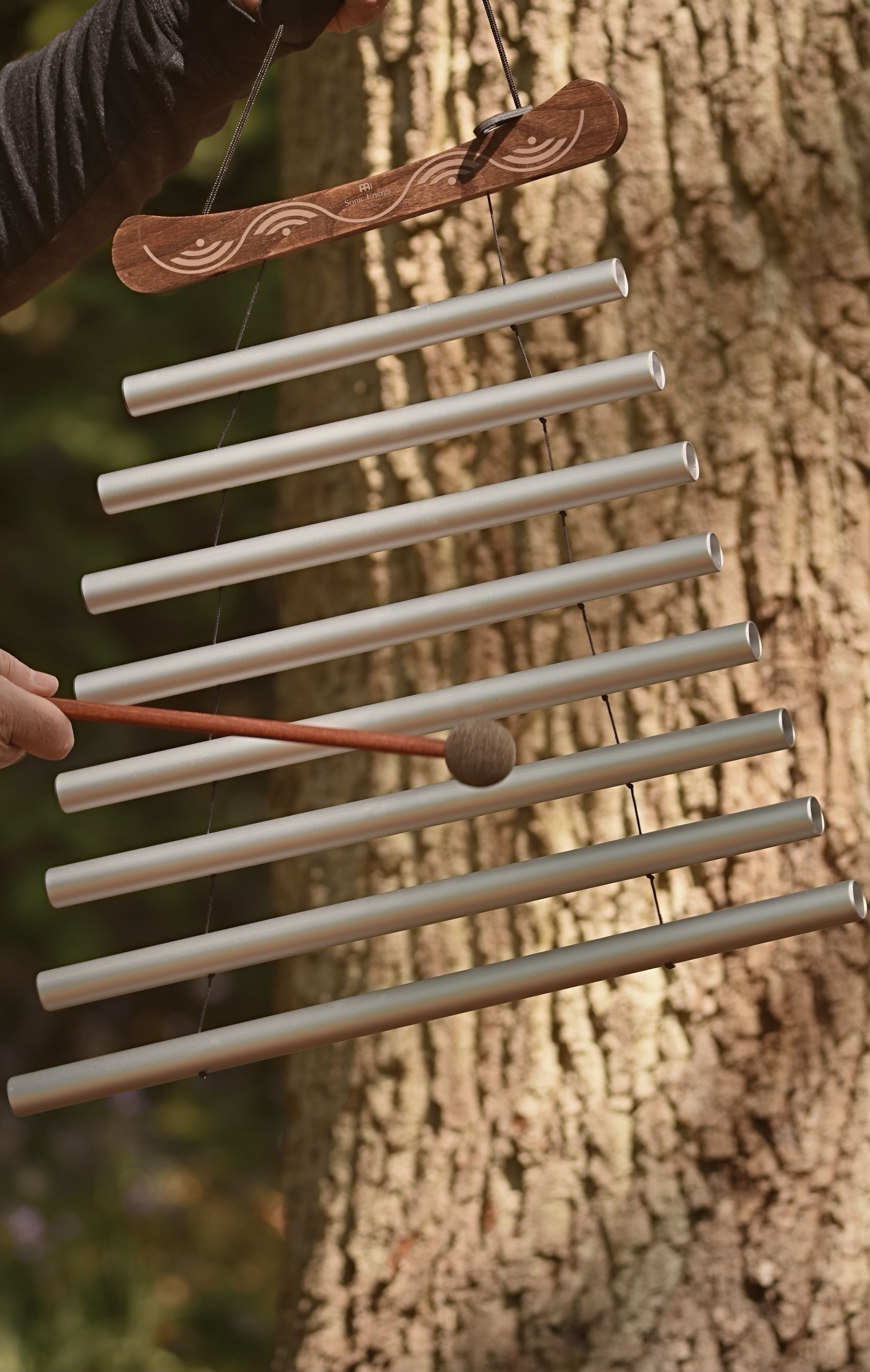 Close-up of a person playing a metal chime xylophone outdoors, with a tree trunk in the background.