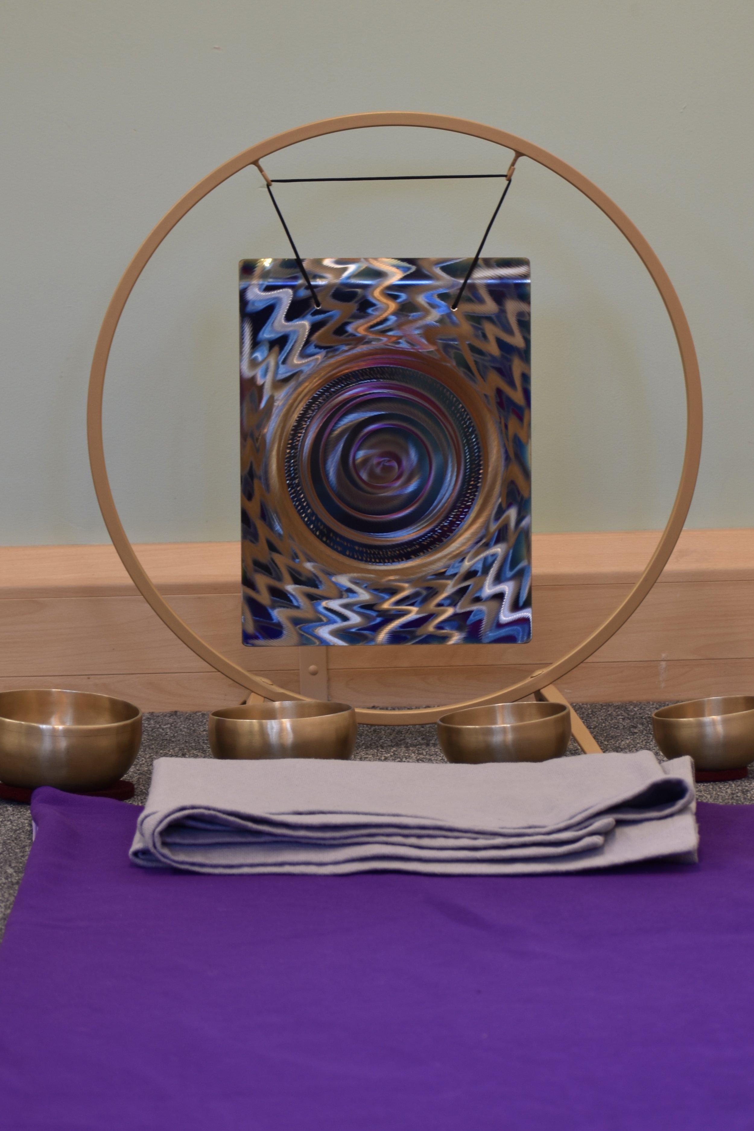 A Tibetan singing bowl setup with four brass bowls, a purple mat, folded gray cloth, and a circular wooden stand holding an abstract patterned mirror with swirling colors.