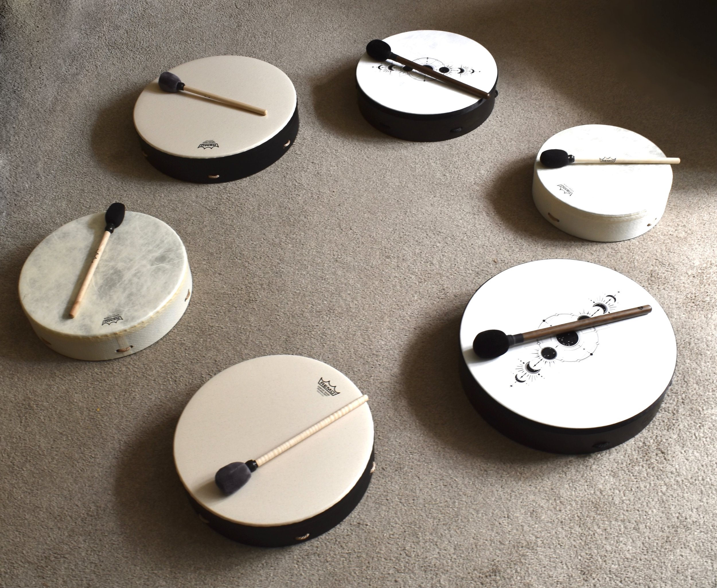 Five percussion drums with mallets on a carpeted floor, arranged in a circle. The drums are of different sizes and colors, with some featuring decorative patterns.