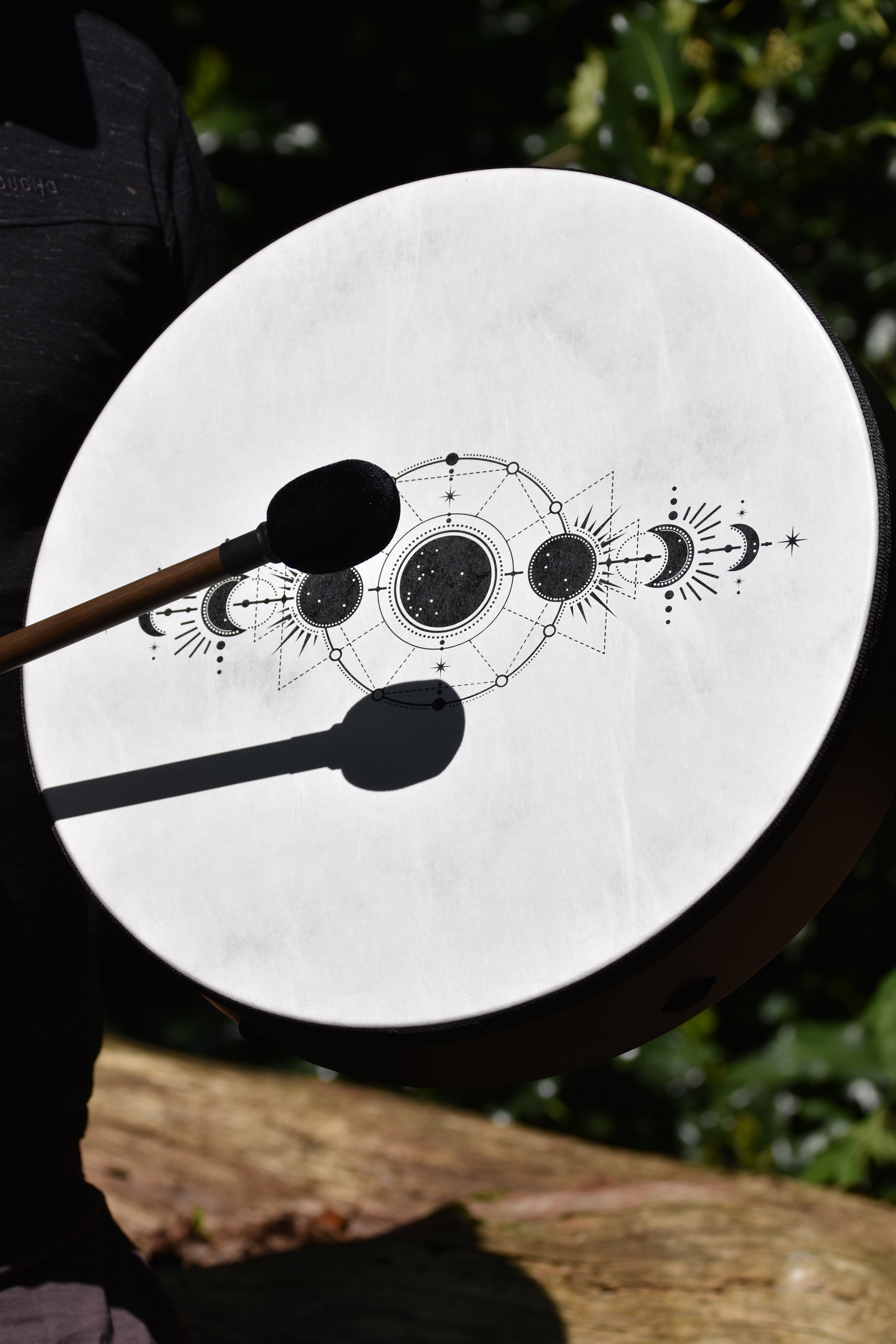 A hand-held drum with celestial-themed designs, including moons and stars, with a black mallet resting on it, outdoors with greenery in the background.
