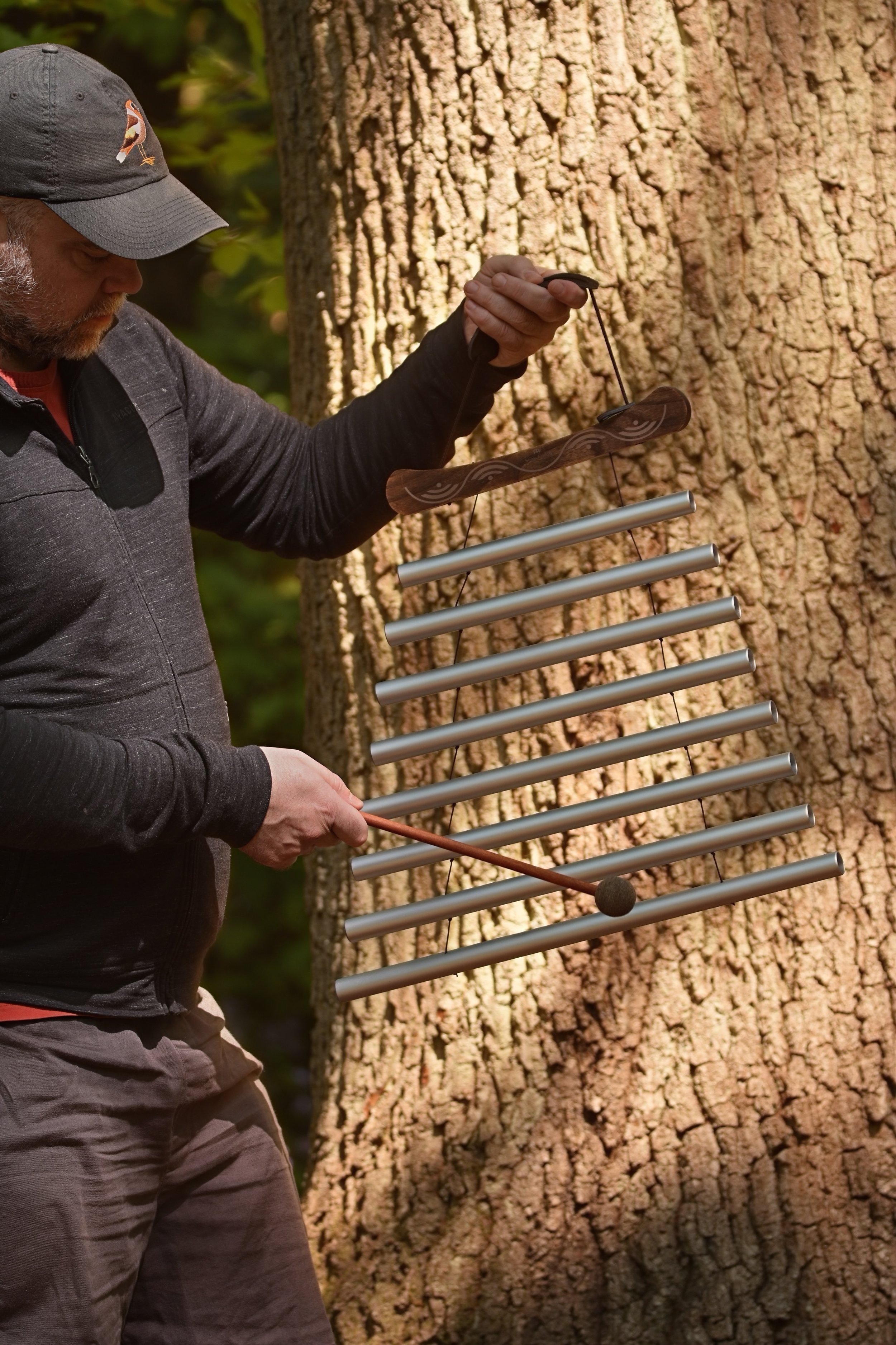 A man standing outdoors playing a metal xylophone-like musical instrument attached to a tree, using mallets.