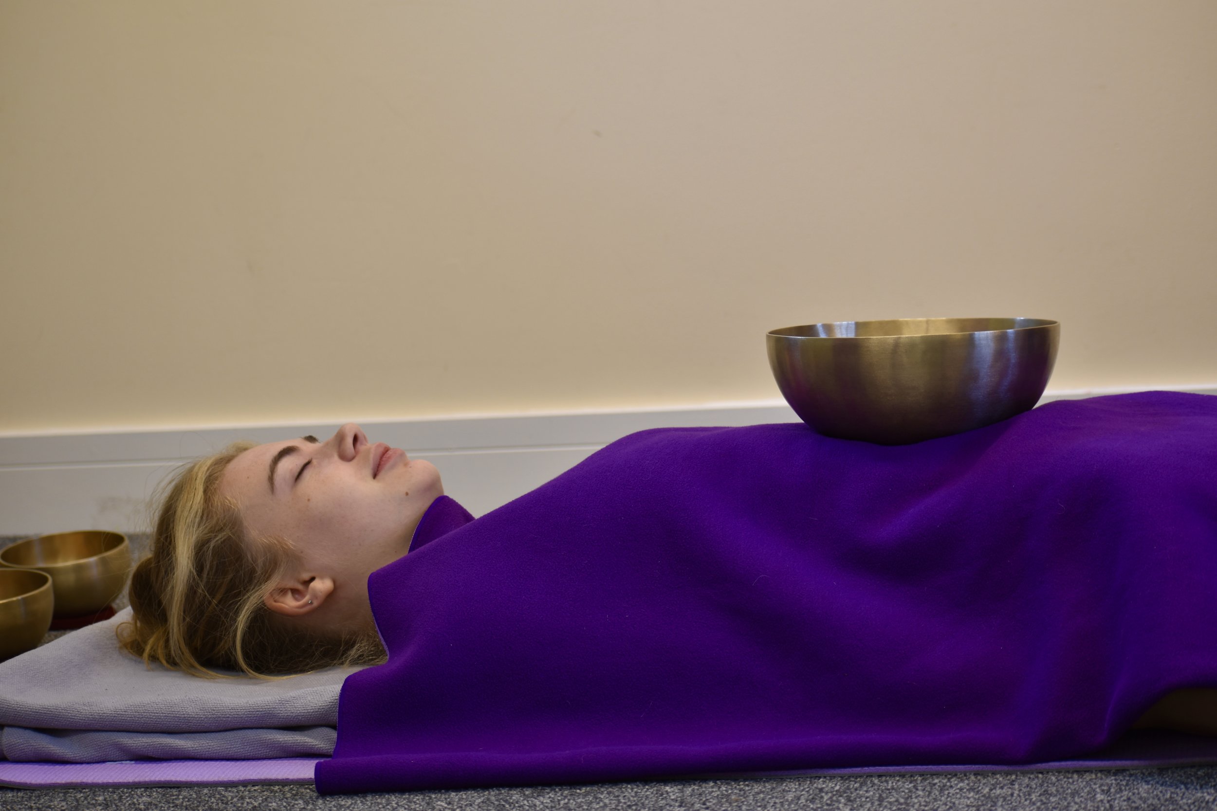 A woman with blonde hair is lying down with her eyes closed. She is covered in a purple blanket, and there are Tibetan singing bowls placed on her chest and near her head, suggesting a sound healing or relaxation session.