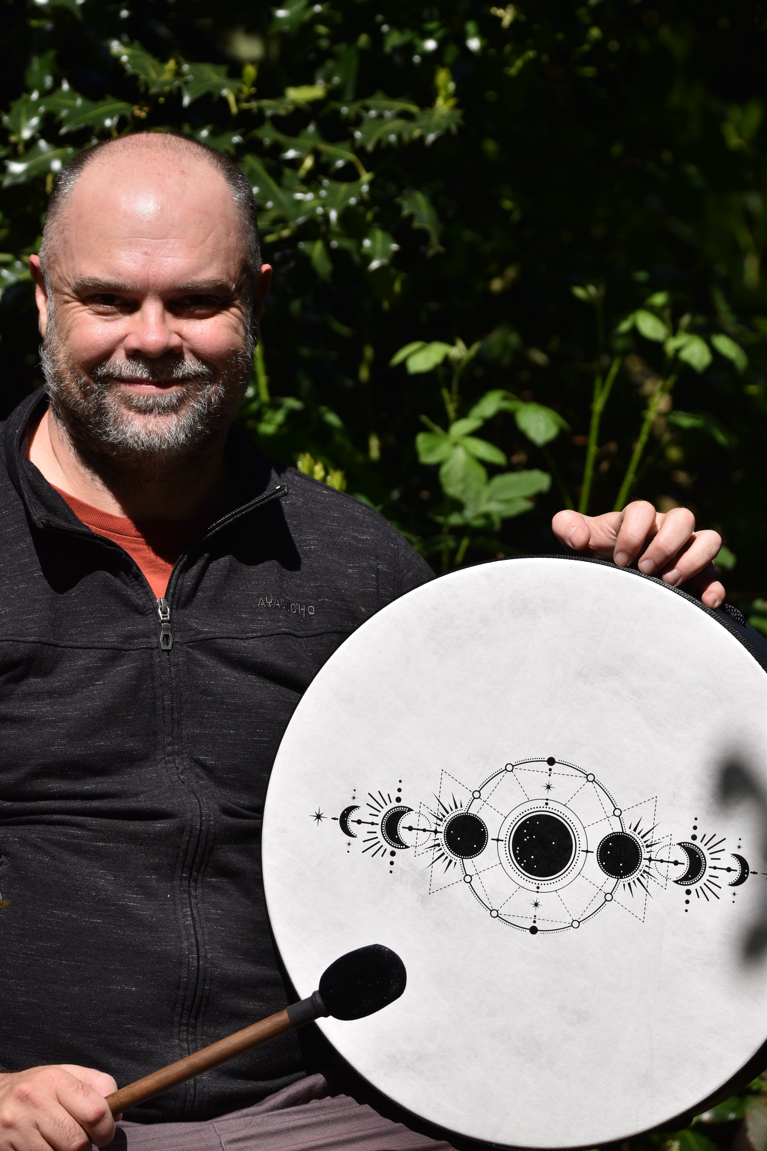 A smiling man with a gray beard and bald head holding a large round drum decorated with celestial and moon phases illustrations, outdoors with green foliage in the background.