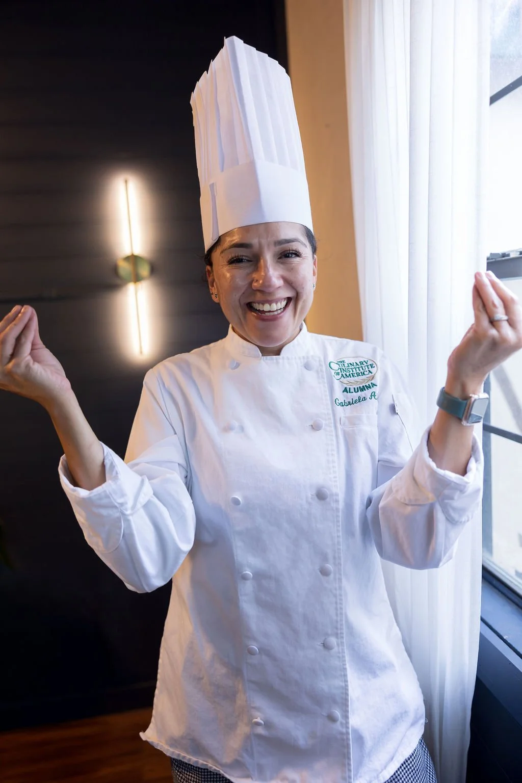 Young woman chef wearing a tall white chef's hat and white chef's coat smiling and making a peace sign with both hands, standing near a window with white curtains.