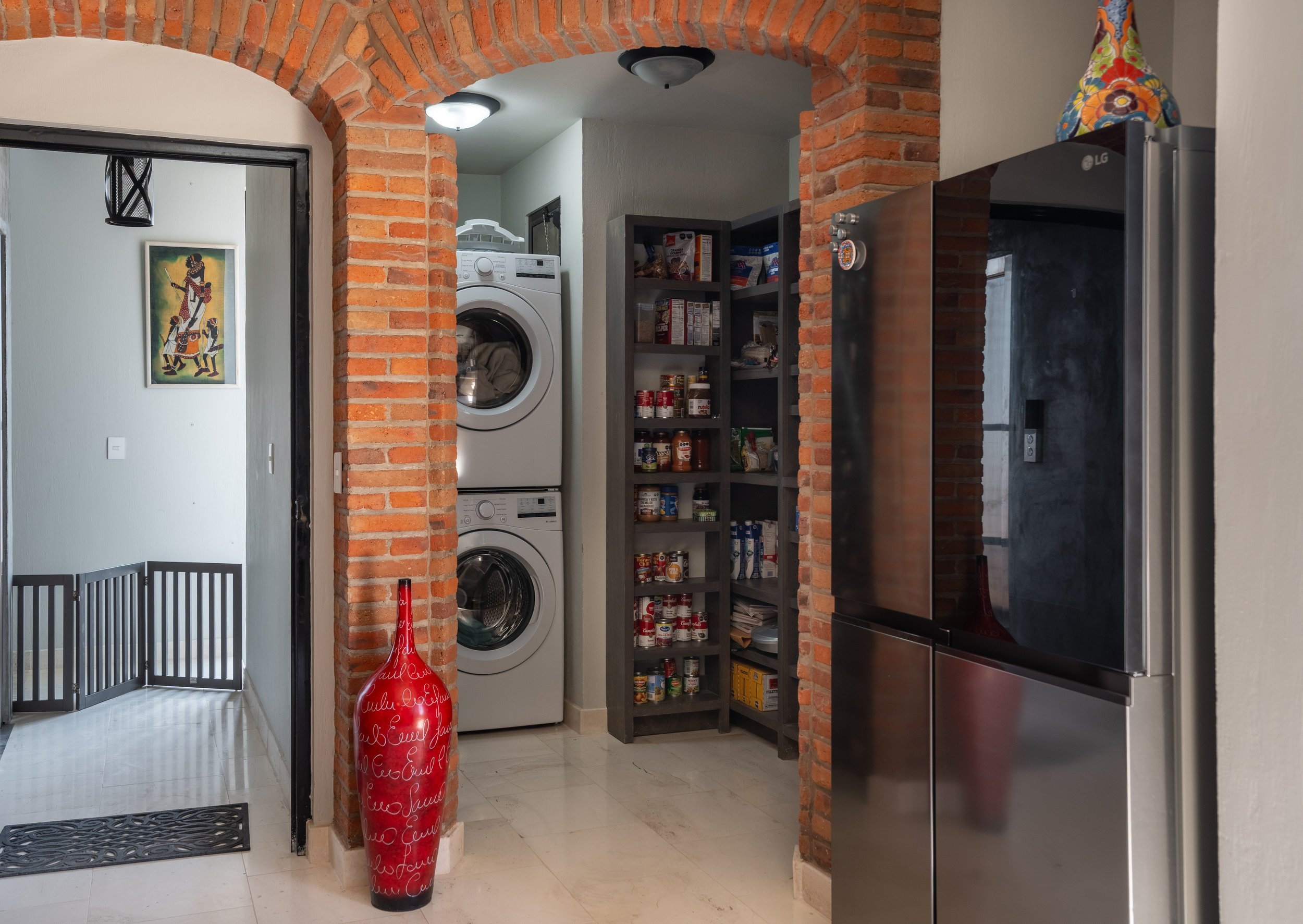 Indoor space with exposed brick walls, a stacked washer and dryer, pantry shelves filled with food items, a stainless steel refrigerator, a colorful vase on top, and a decorative red bottle.