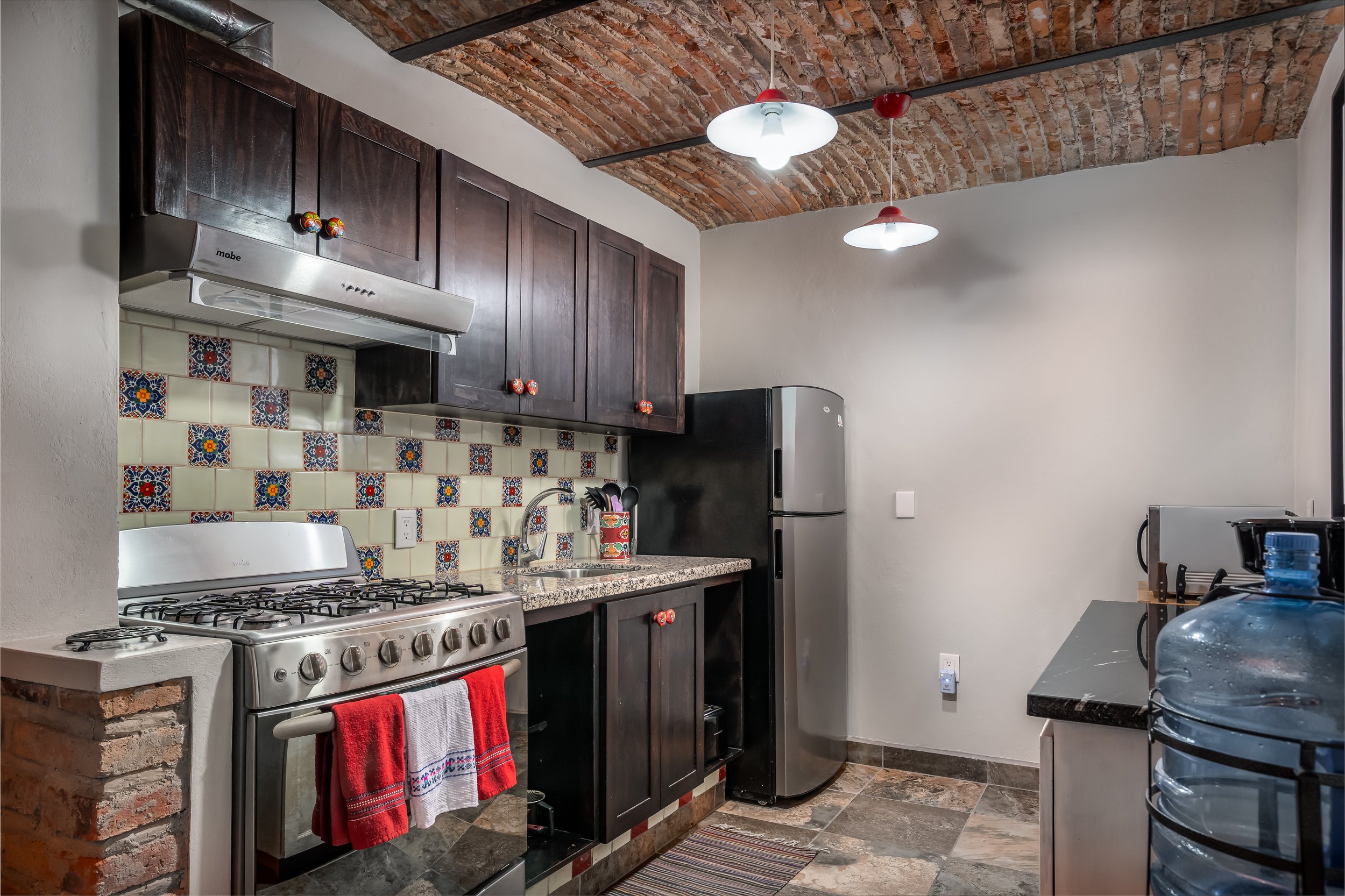Modern kitchen with brick ceiling, stainless steel appliances, dark cabinets, colorful tile backsplash, red and white towel on oven, black refrigerator, and water cooler.