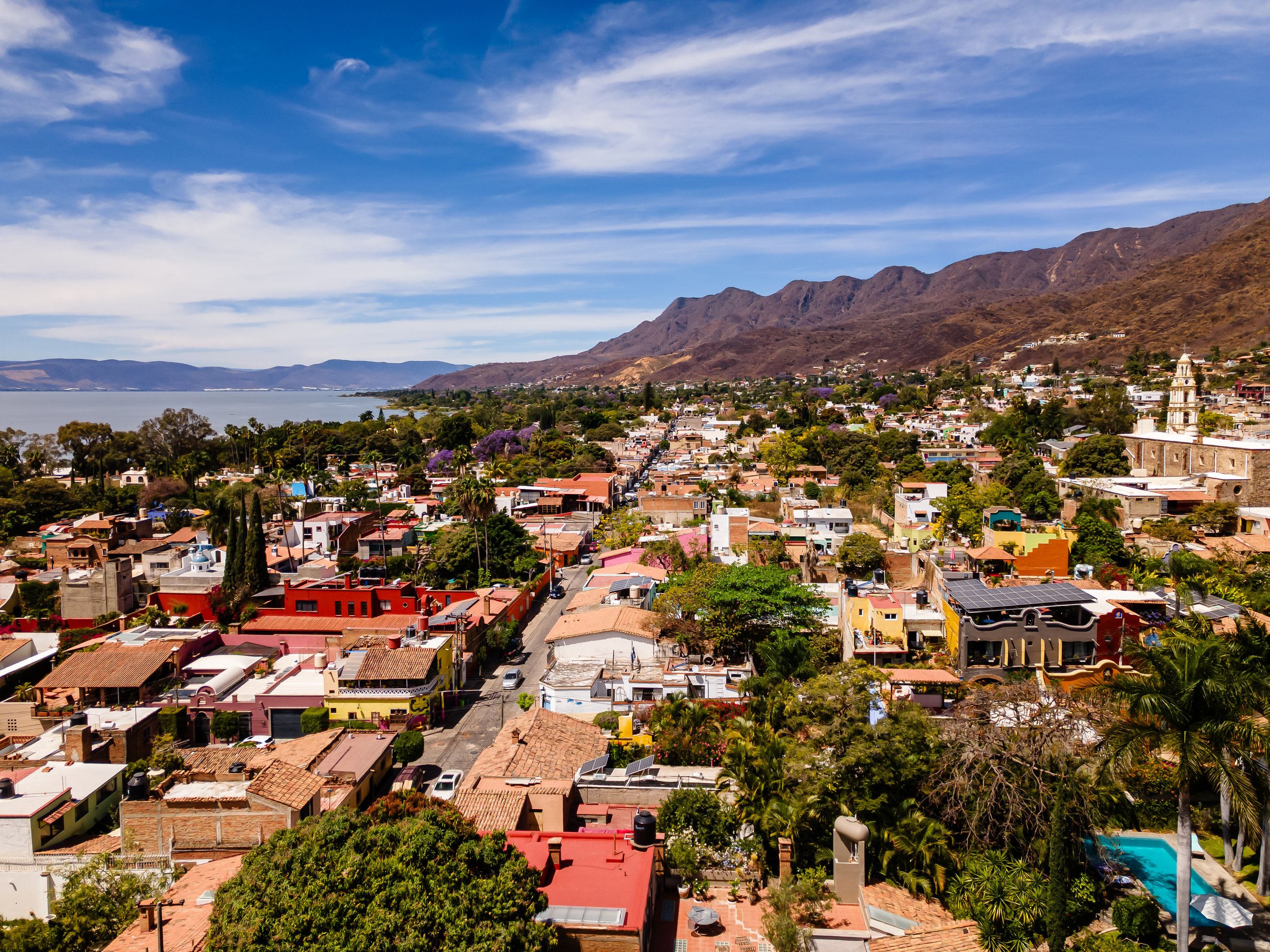 Ajijic rental opportunity. Investment property. Aerial view of a colorful town with a mix of houses and trees, surrounded by mountains and a large body of water under a blue sky with white clouds.