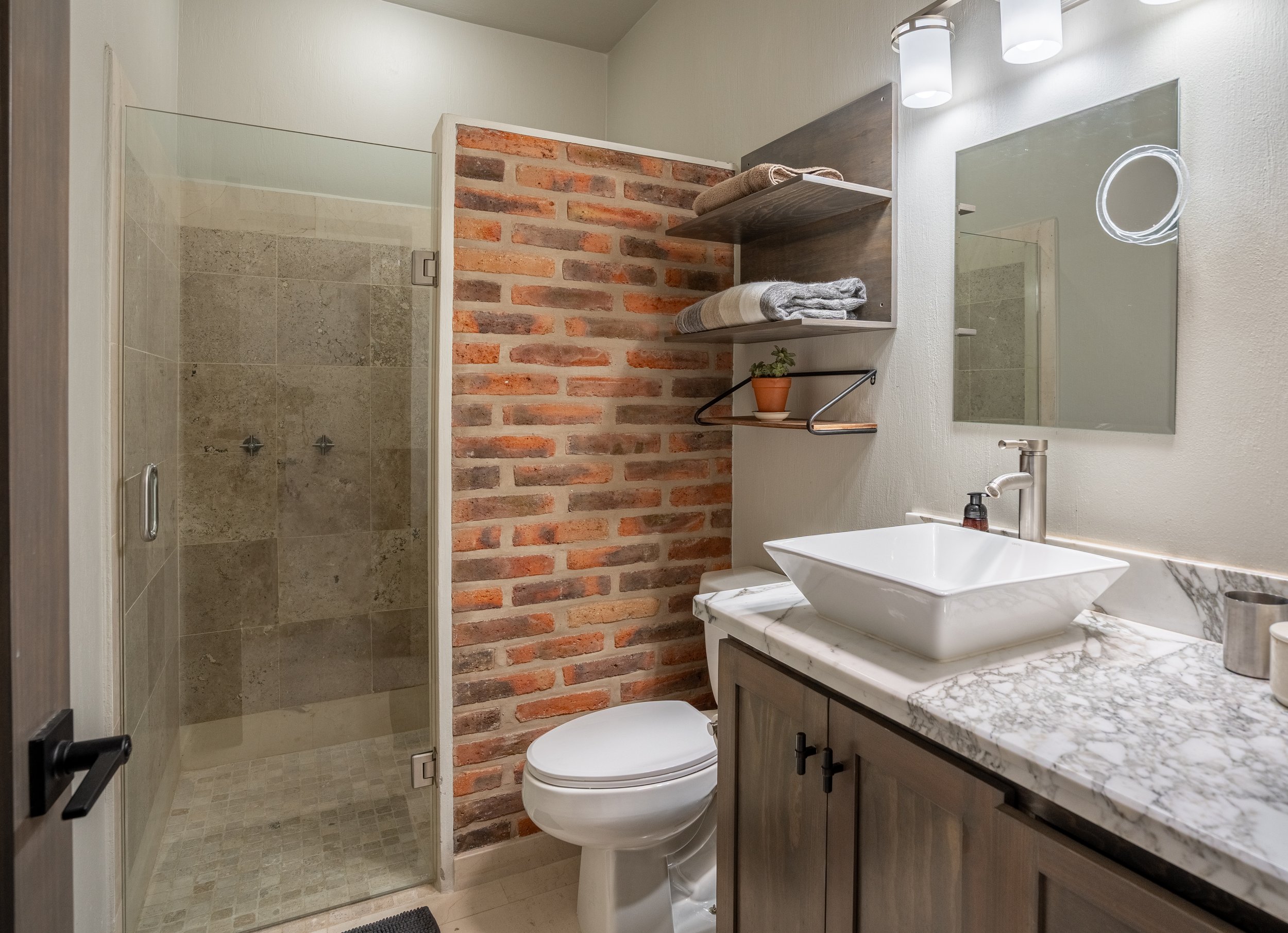 Modern bathroom with brick wall, glass shower, vessel sink, and marble countertop.