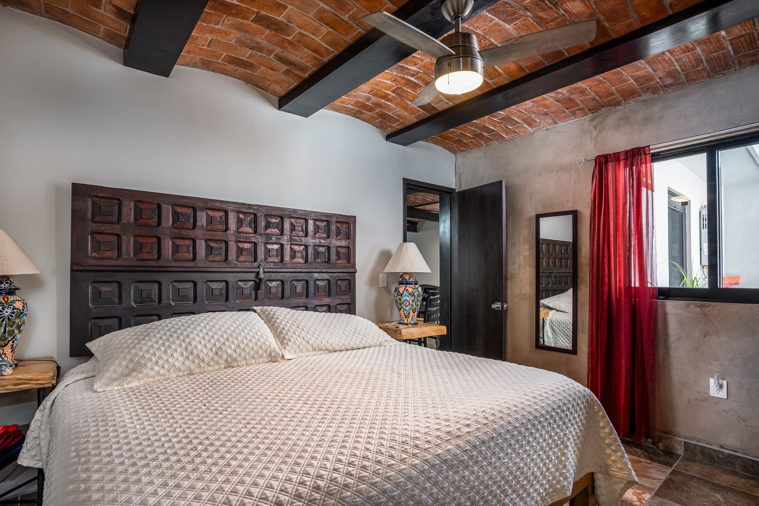 Modern bedroom with brick ceiling, wooden headboard, white bedding, colorful lamp, and red curtains.