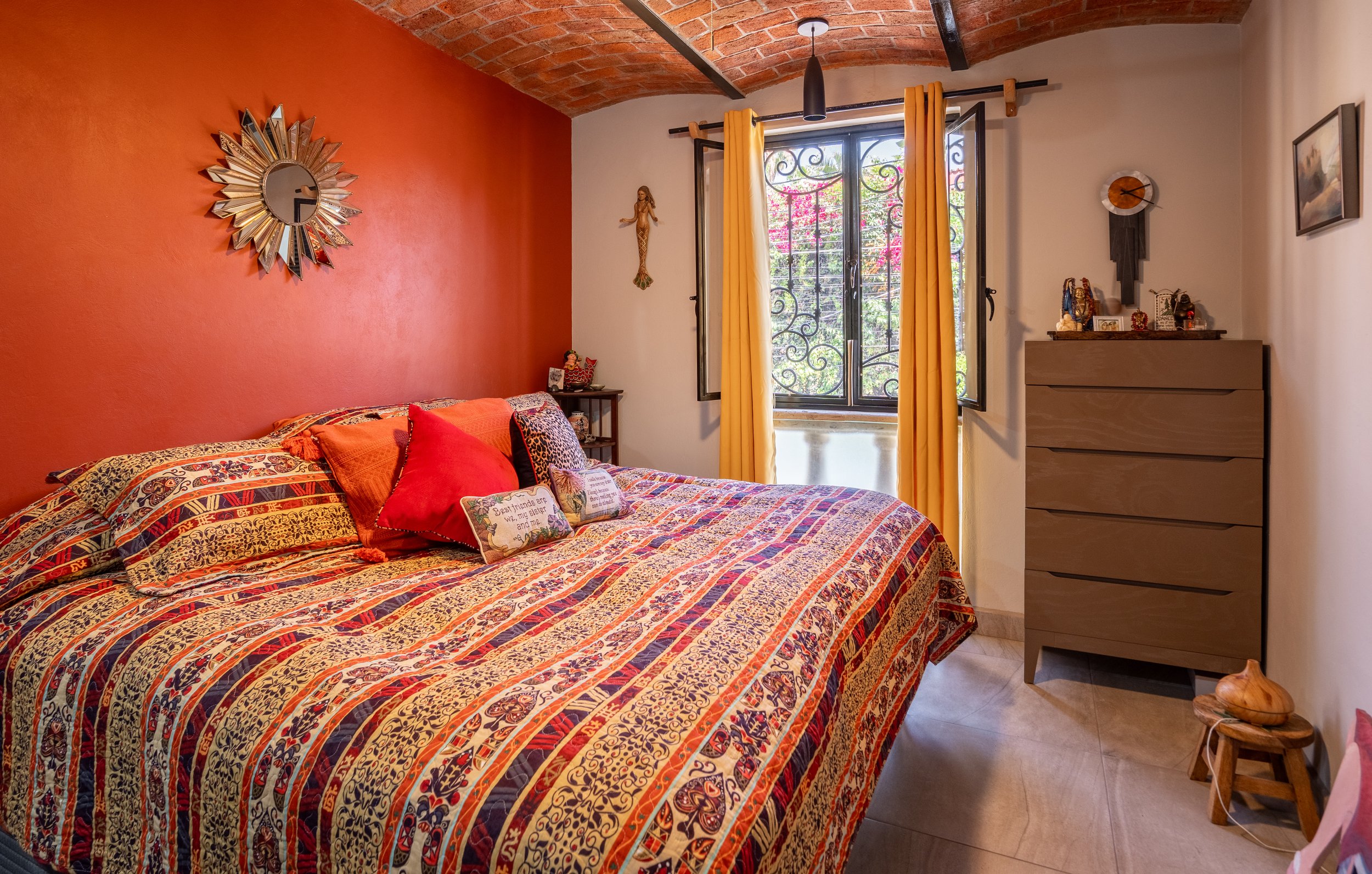 Cozy bedroom with an orange accent wall, featuring a bed with colorful patterned bedding and decorative pillows. A sunburst mirror hangs on the wall above the bed. A window with yellow curtains allows natural light in. Beside the bed, a small nightst