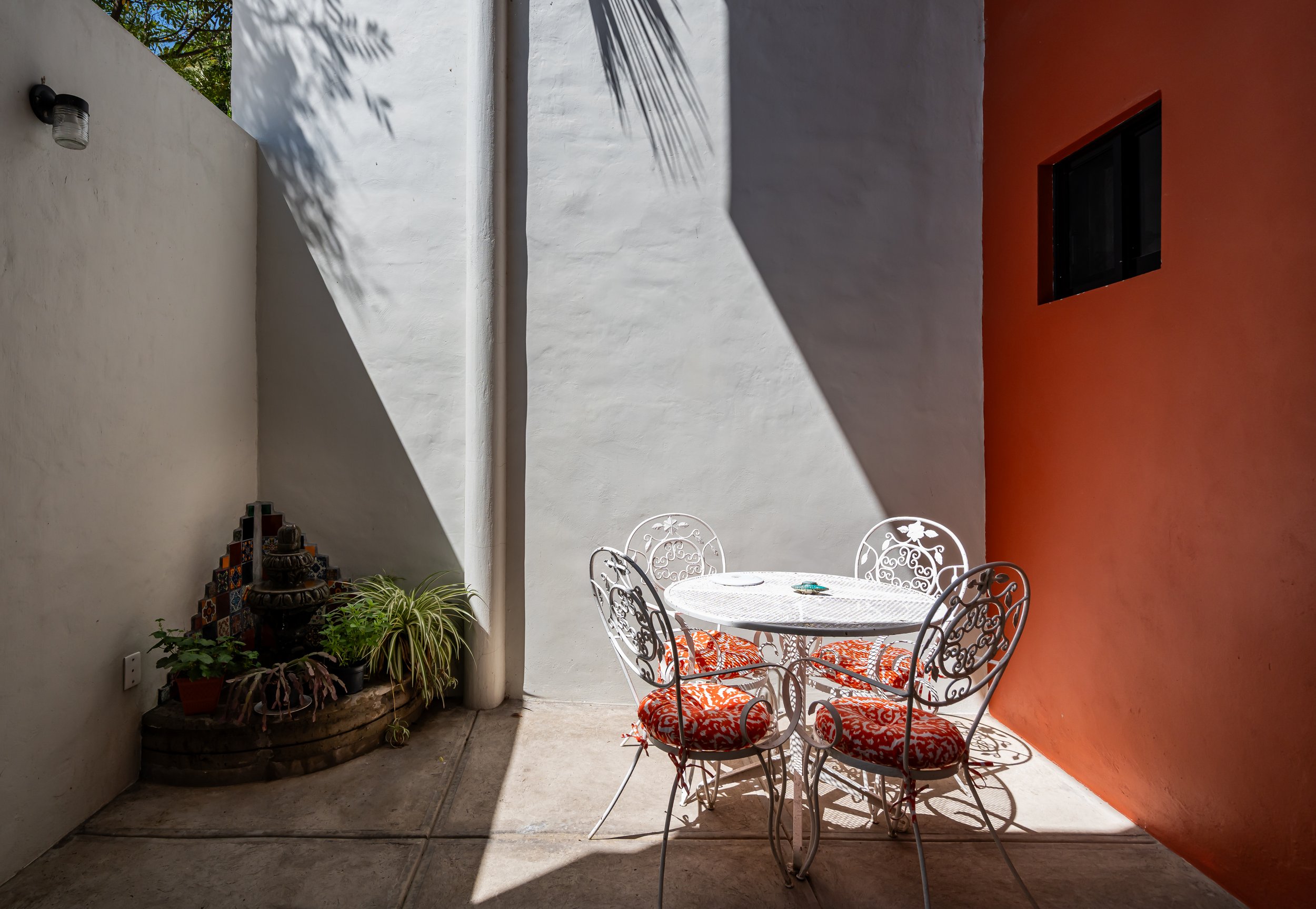 Outdoor patio with white iron furniture set, featuring four chairs with red and white patterned cushions around a white table. A small corner fountain and plants are on the left. Sunlight casts shadows on the white and orange walls.