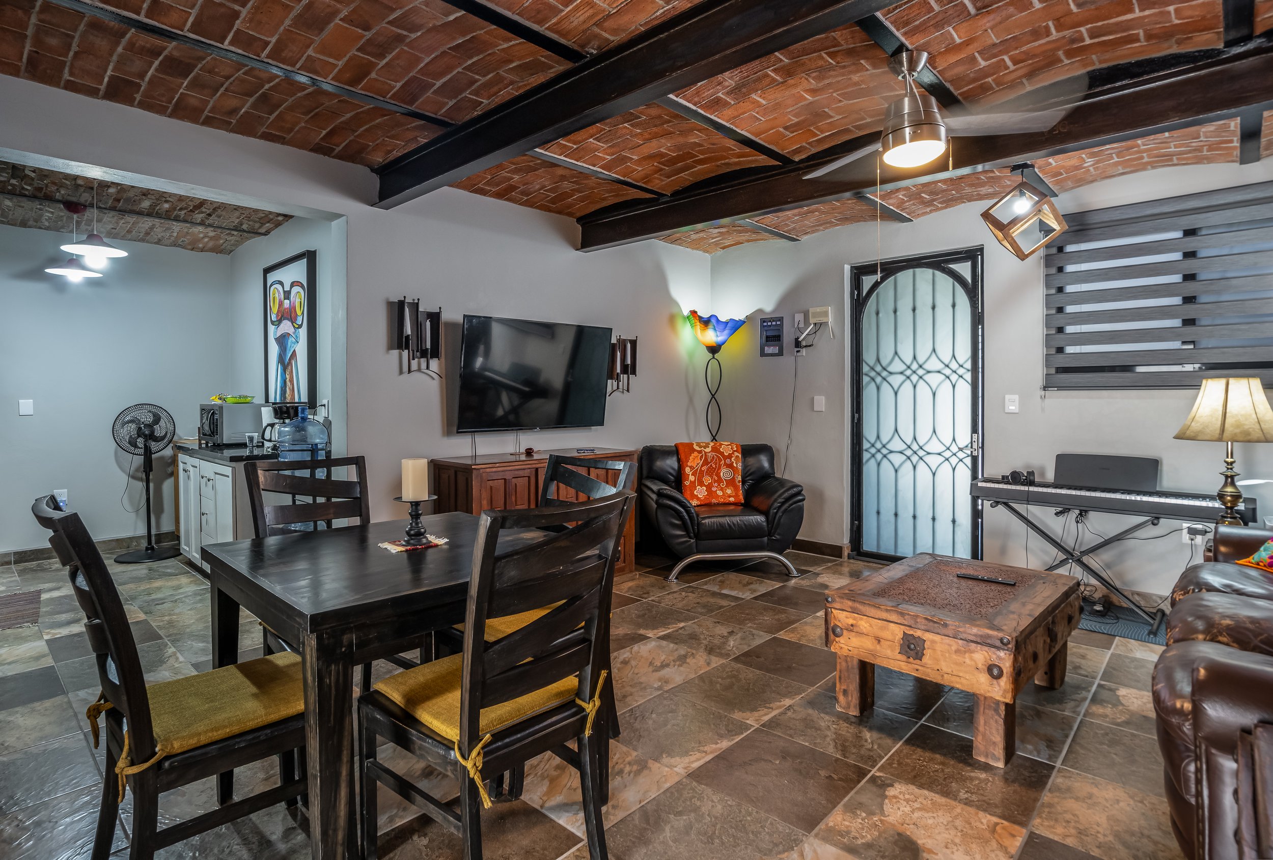 Cozy living room with brick ceiling, black dining table with chairs, TV on wall, leather armchairs, colorful lamp, wooden coffee table, and electric piano.