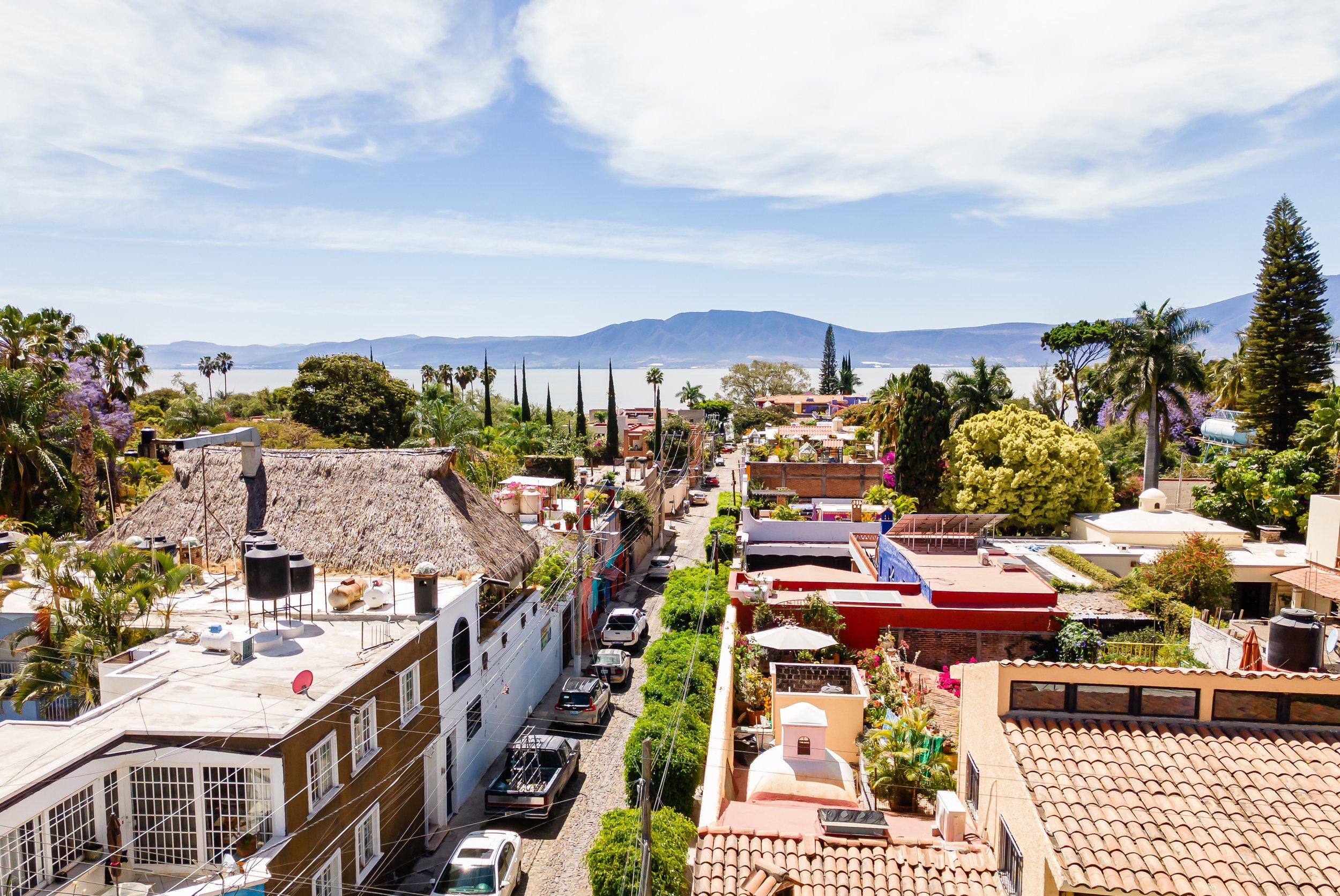 Scenic view of Ajijic with tiled rooftops, narrow streets, and lush greenery, overlooking Lake Chapala and distant mountains under a blue sky.