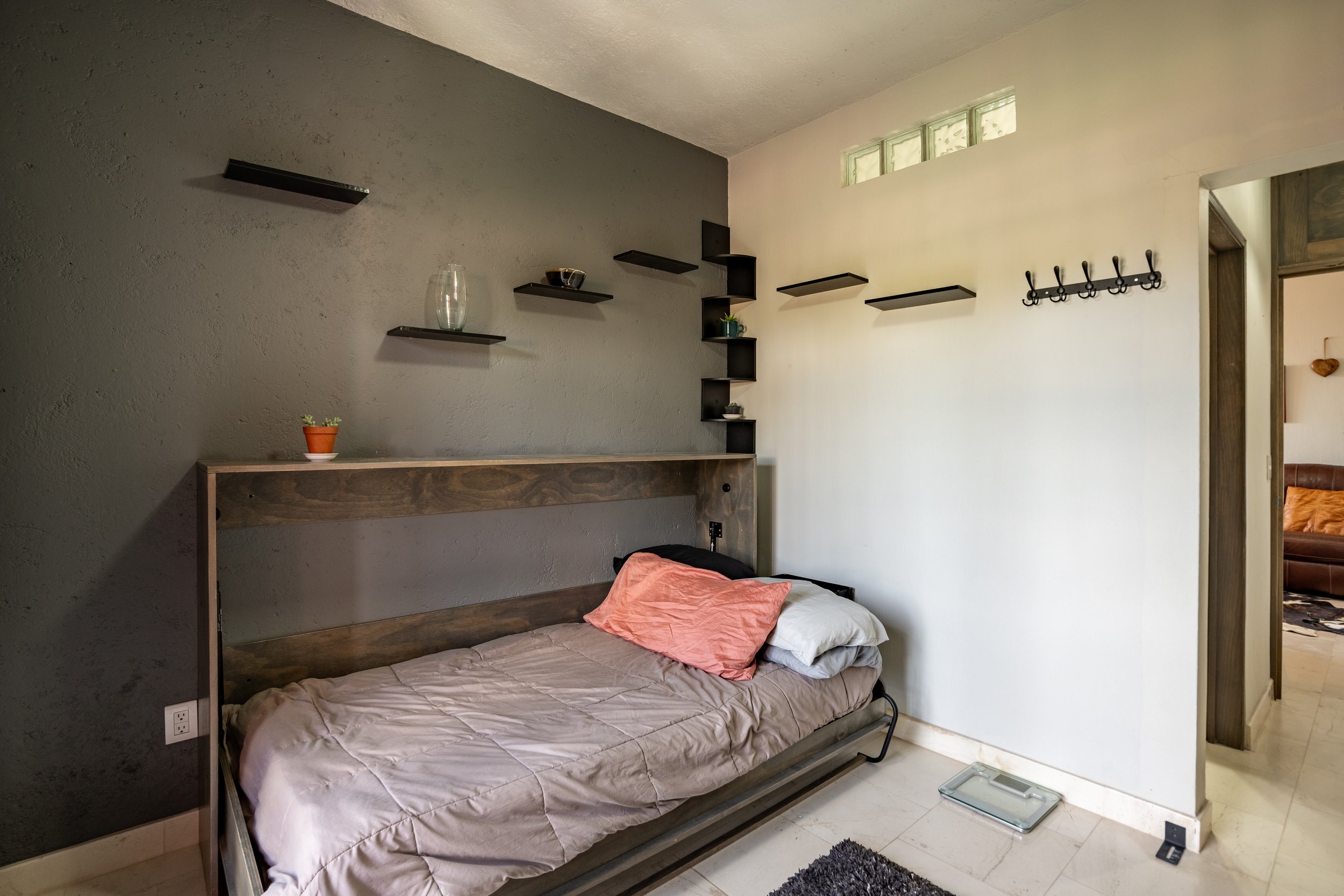Minimalist bedroom with a single bed, gray wall, floating shelves, and a coat rack.