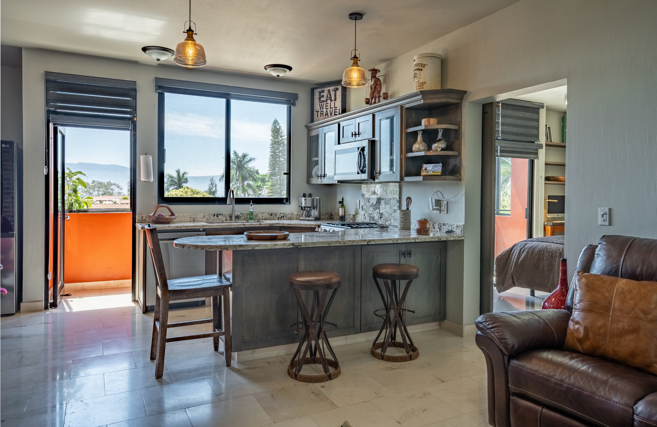 Modern kitchen with a breakfast bar, pendant lights, wooden cabinets, bar stools, and a view of an outdoor balcony with palm trees.