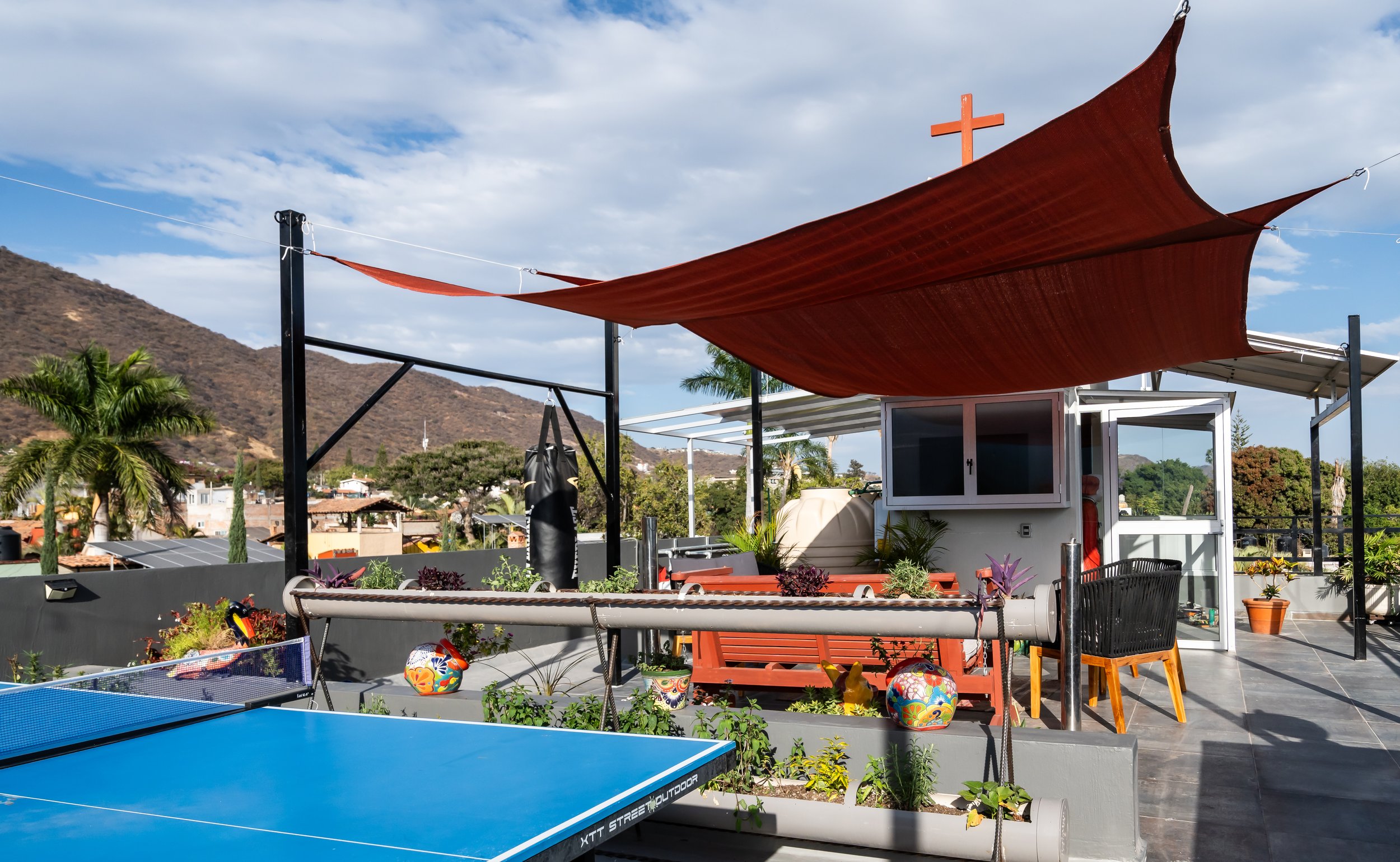 Ajijic rental opportunity. Investment property. Rooftop terrace with a blue ping pong table and potted plants under a red sunshade. Decor includes a punching bag, colorful ceramic vases, and a wooden bench.