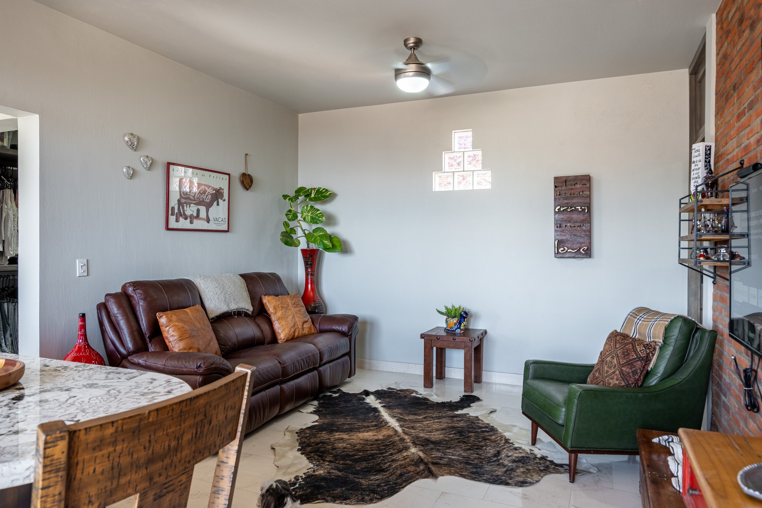 Cozy living room with brown leather sofa, green armchair, cowhide rug, wall art, potted plant, and ceiling fan.