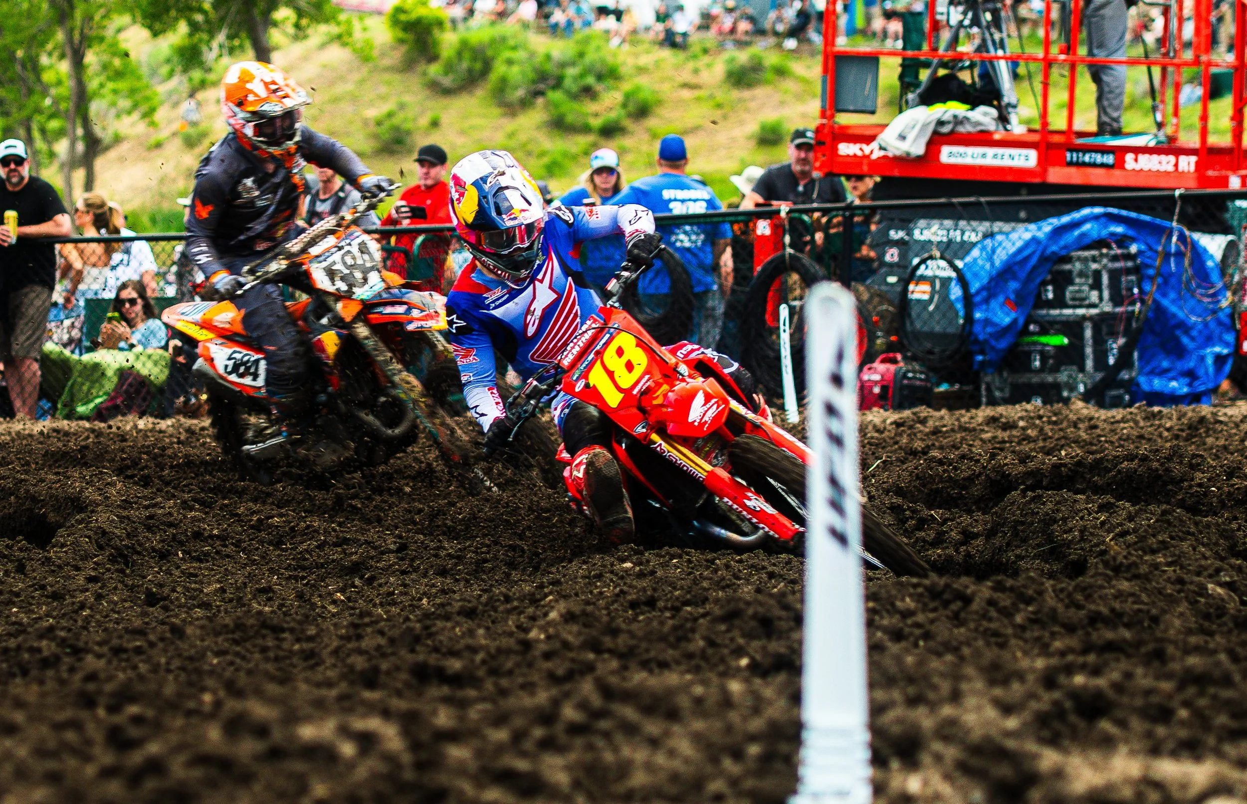 Two motocross riders racing on a dirt track with a crowd of spectators in the background.