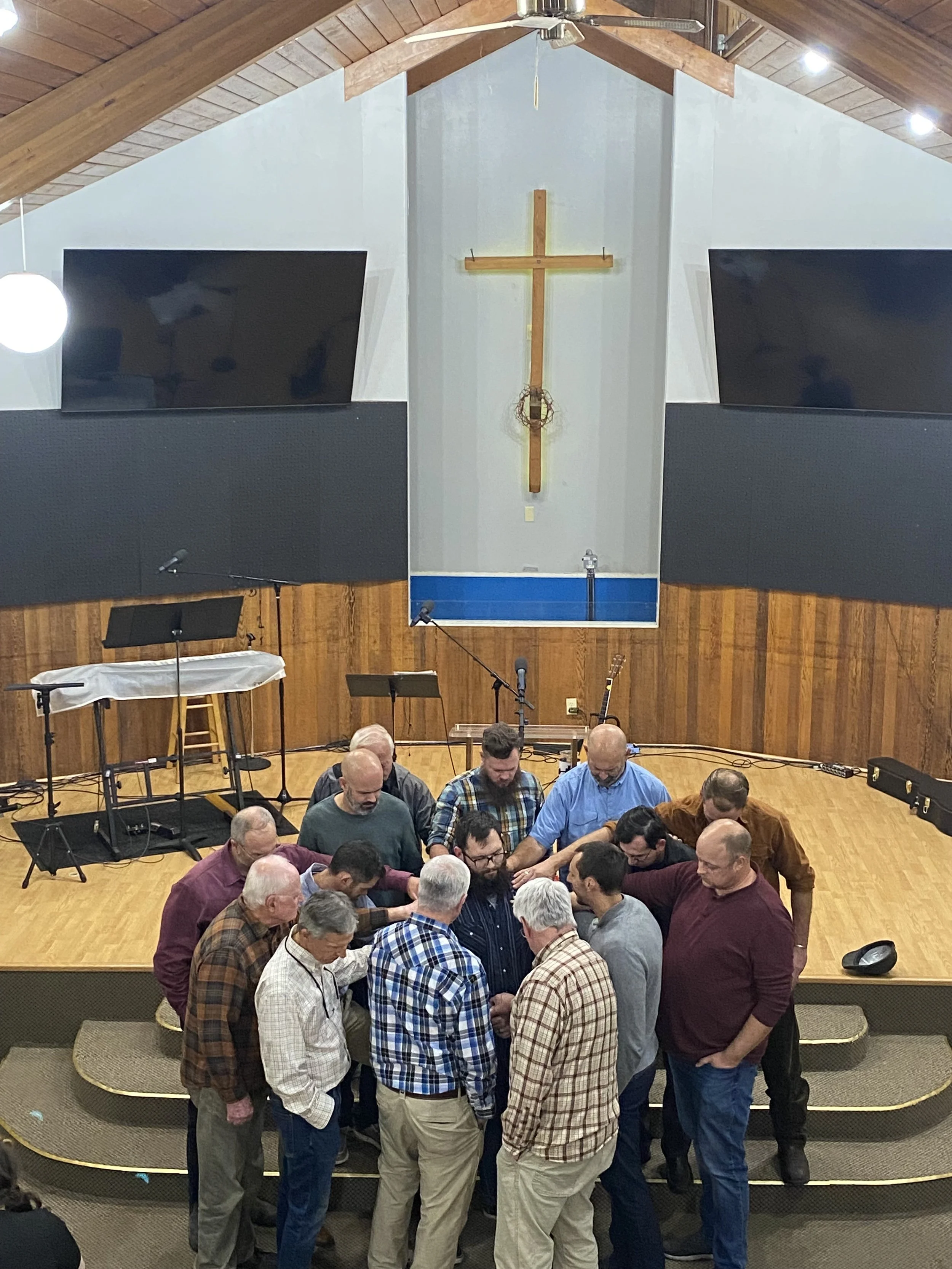 A group of people gathered on a stage inside a Baptist church, with a large wooden cross hanging on the wall behind them during an ordination.
