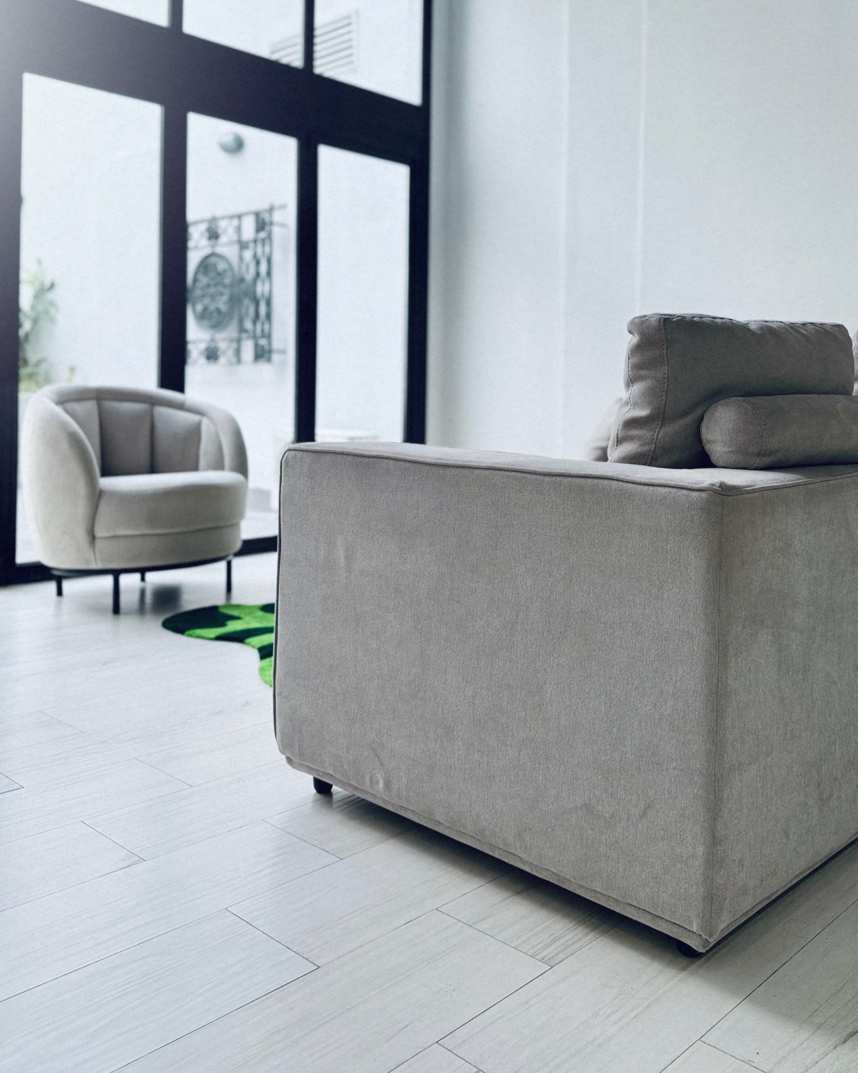 Salón moderno con sillón gris claro, una silla blanca y ventanas grandes con marco negro. El suelo es de baldosas claras y hay una pared blanca y una ventana en el fondo.